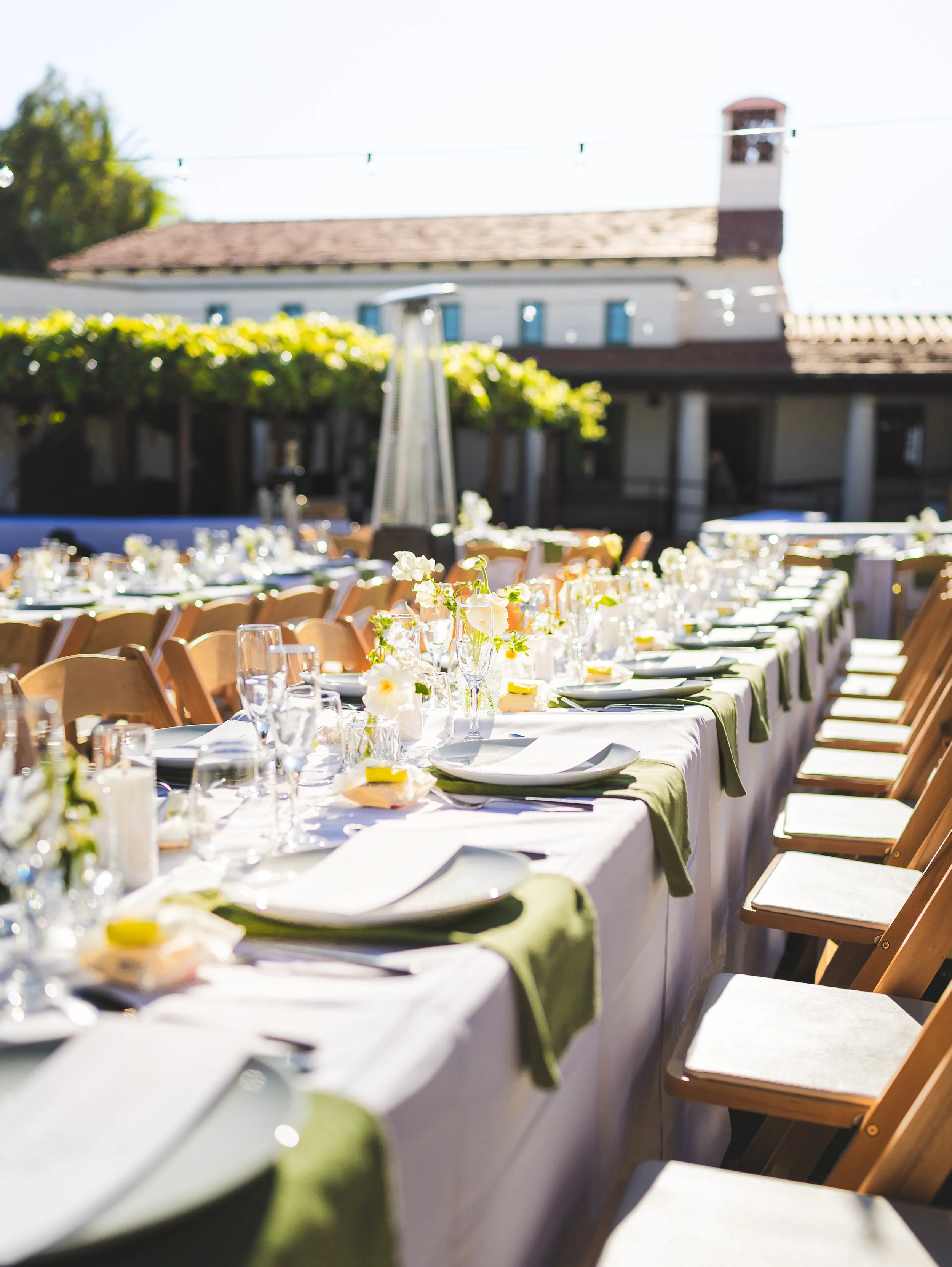 Long outdoor banquet table set for a celebration with white tablecloths, green napkins, floral centerpieces, and glassware, in a sunny garden with a house in the background.