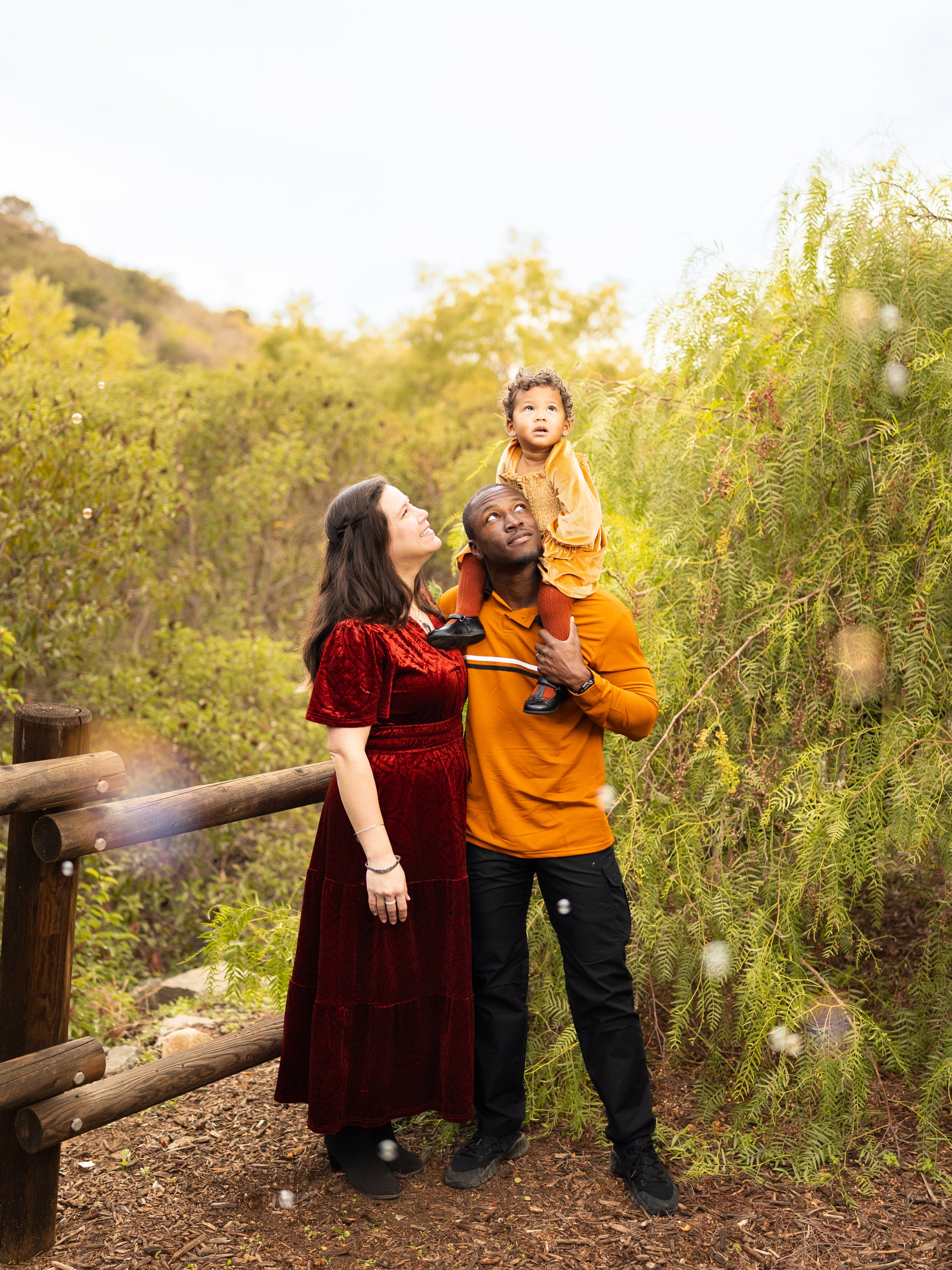 A family of three standing outdoors on a trail in a wooded area during autumn, with the man carrying a young girl on his shoulders and the woman standing beside him.