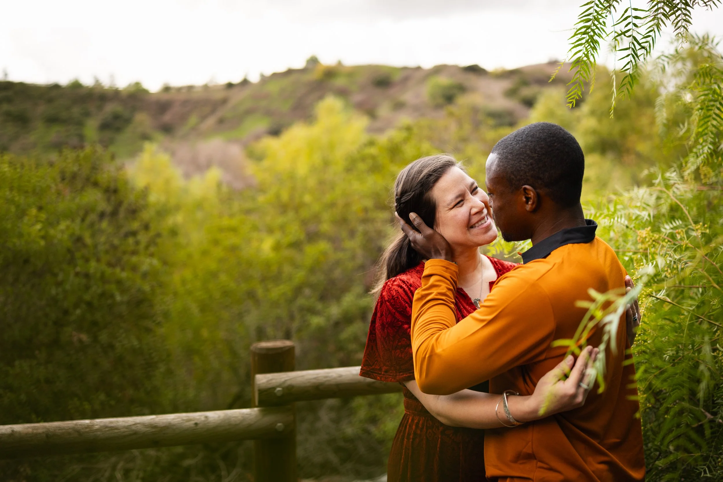 A couple embraces outdoors on a trail surrounded by green foliage, with hills in the background, as they smile and look into each other's eyes.