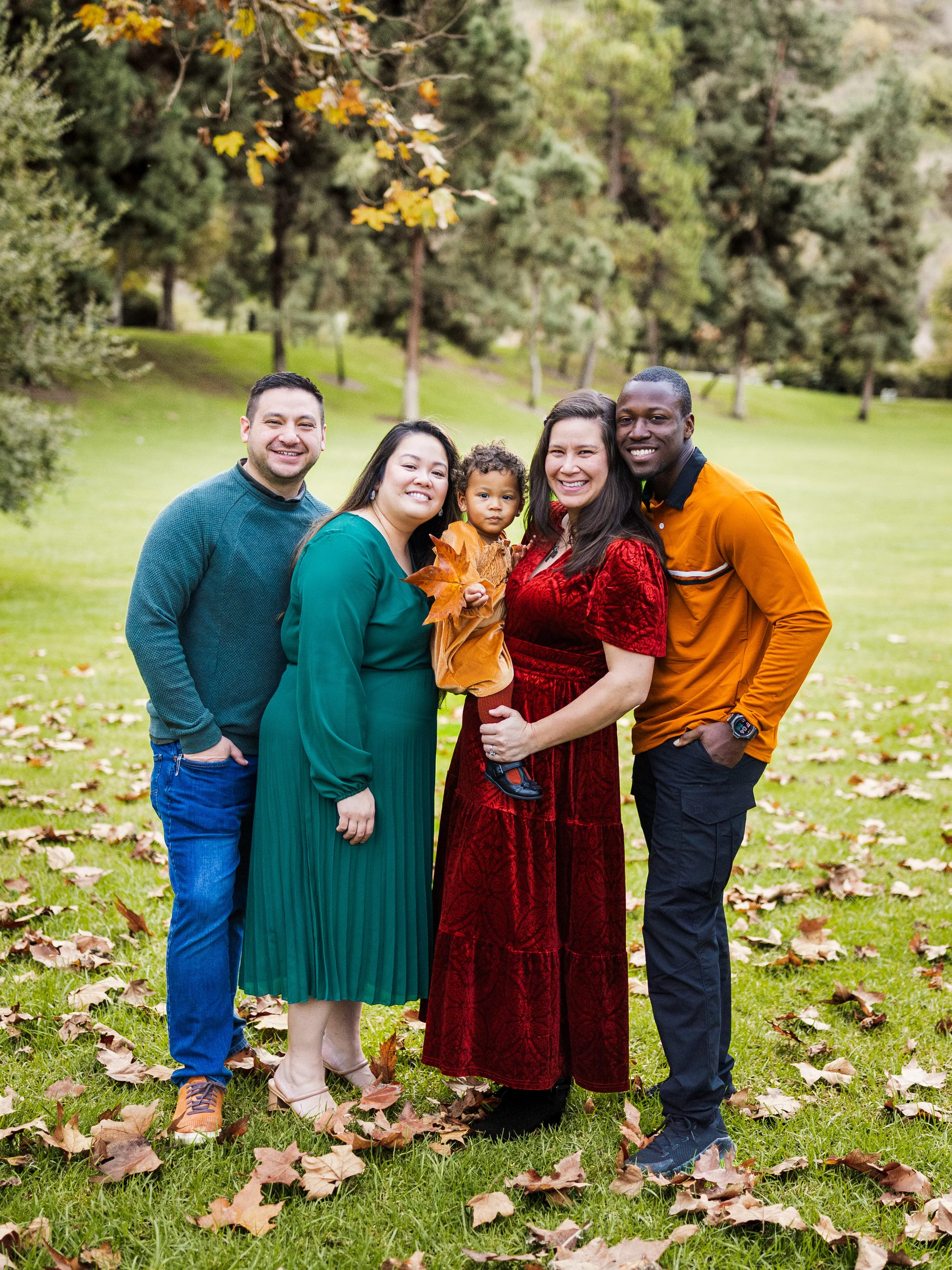 Group of five people, including a woman holding a small child, standing on a grassy park with autumn leaves and trees in the background, smiling at the camera.