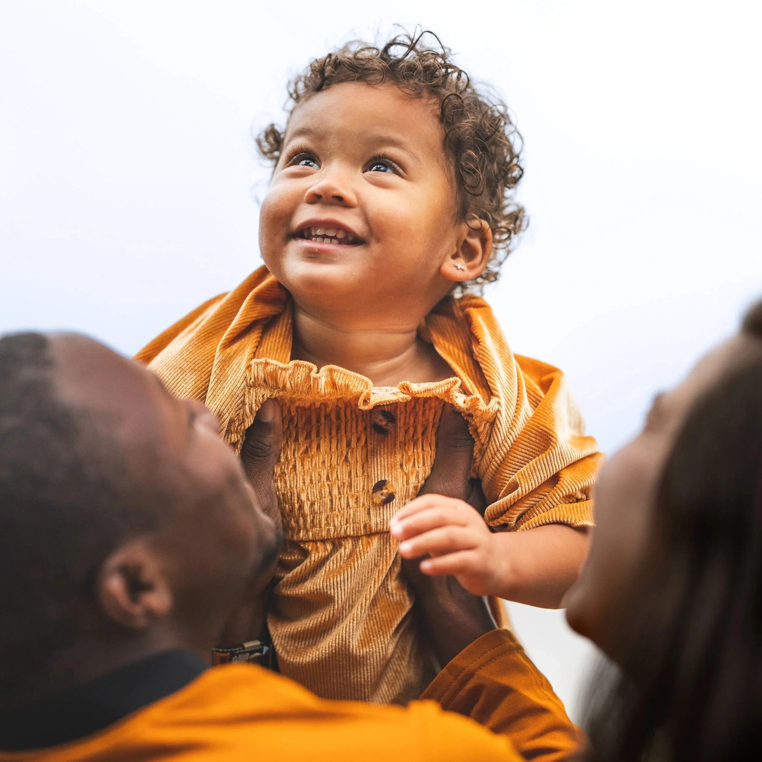 A young child with curly hair smiling while being held up by two adults, with only parts of their faces visible, against a bright sky background.