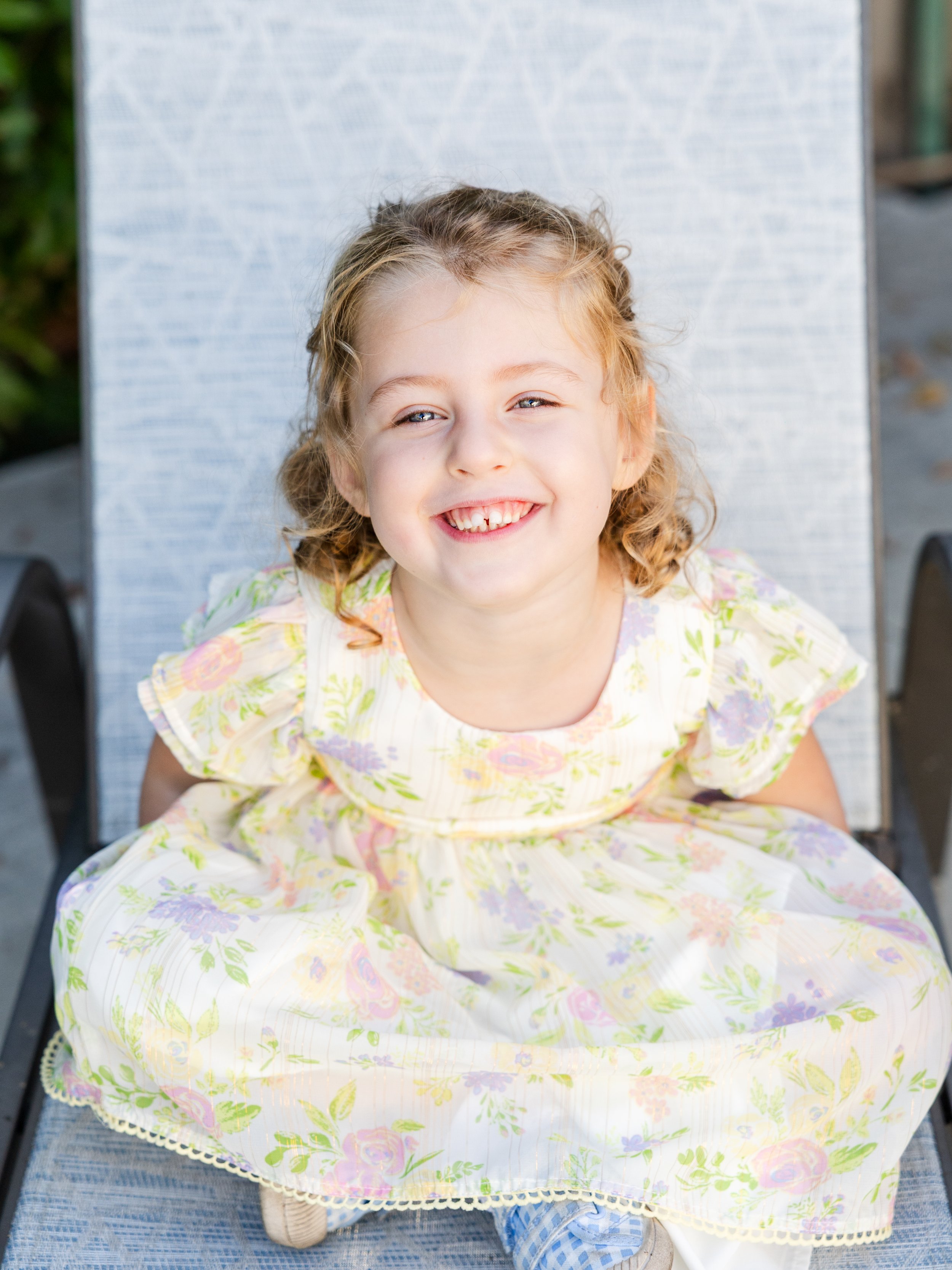 A young girl with curly blonde hair and blue eyes sitting on a lounge chair outdoors, smiling at the camera, wearing a floral dress.
