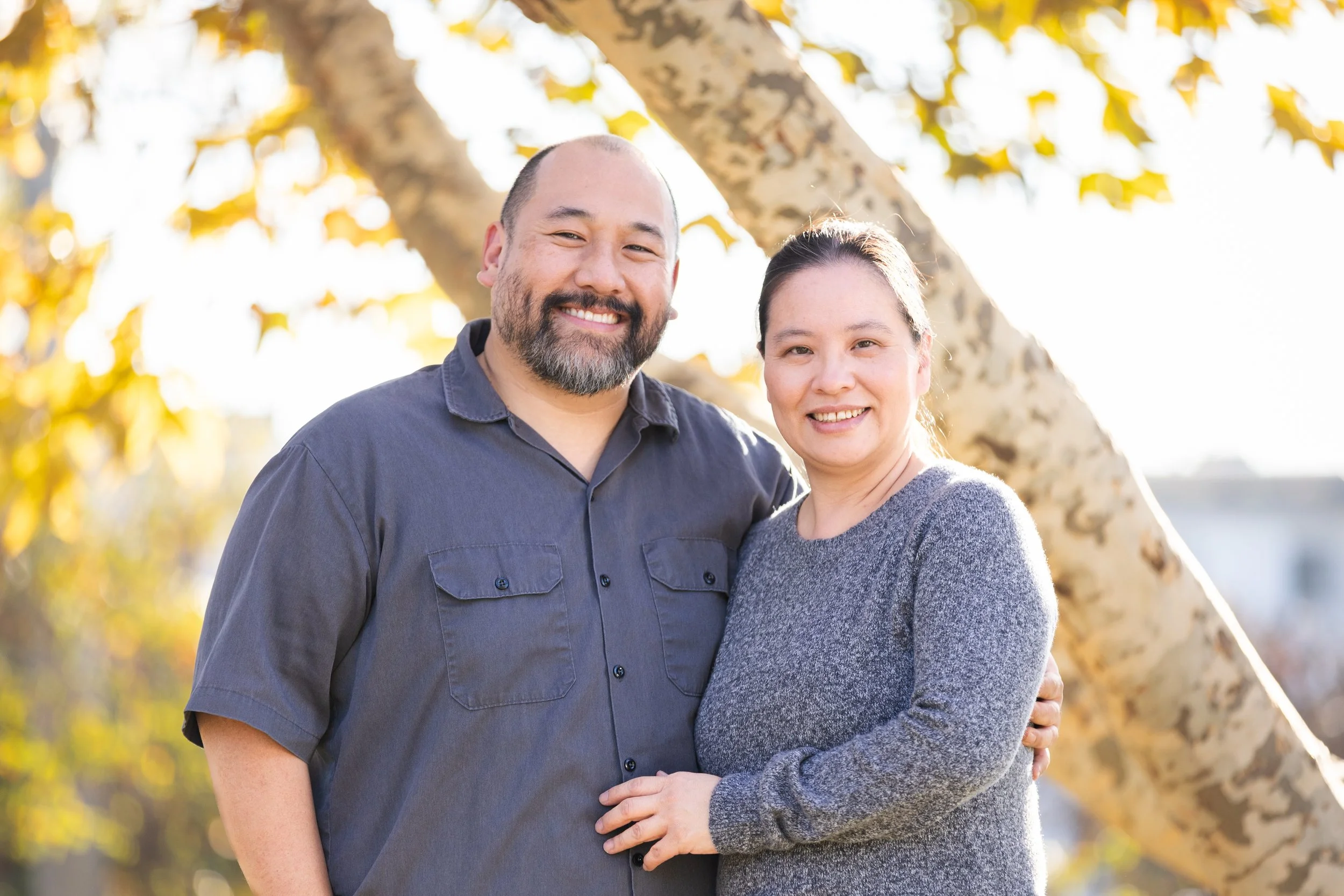 A smiling couple, a man with a beard and a woman, standing outdoors in front of a tree during fall, with sunlight highlighting their faces.