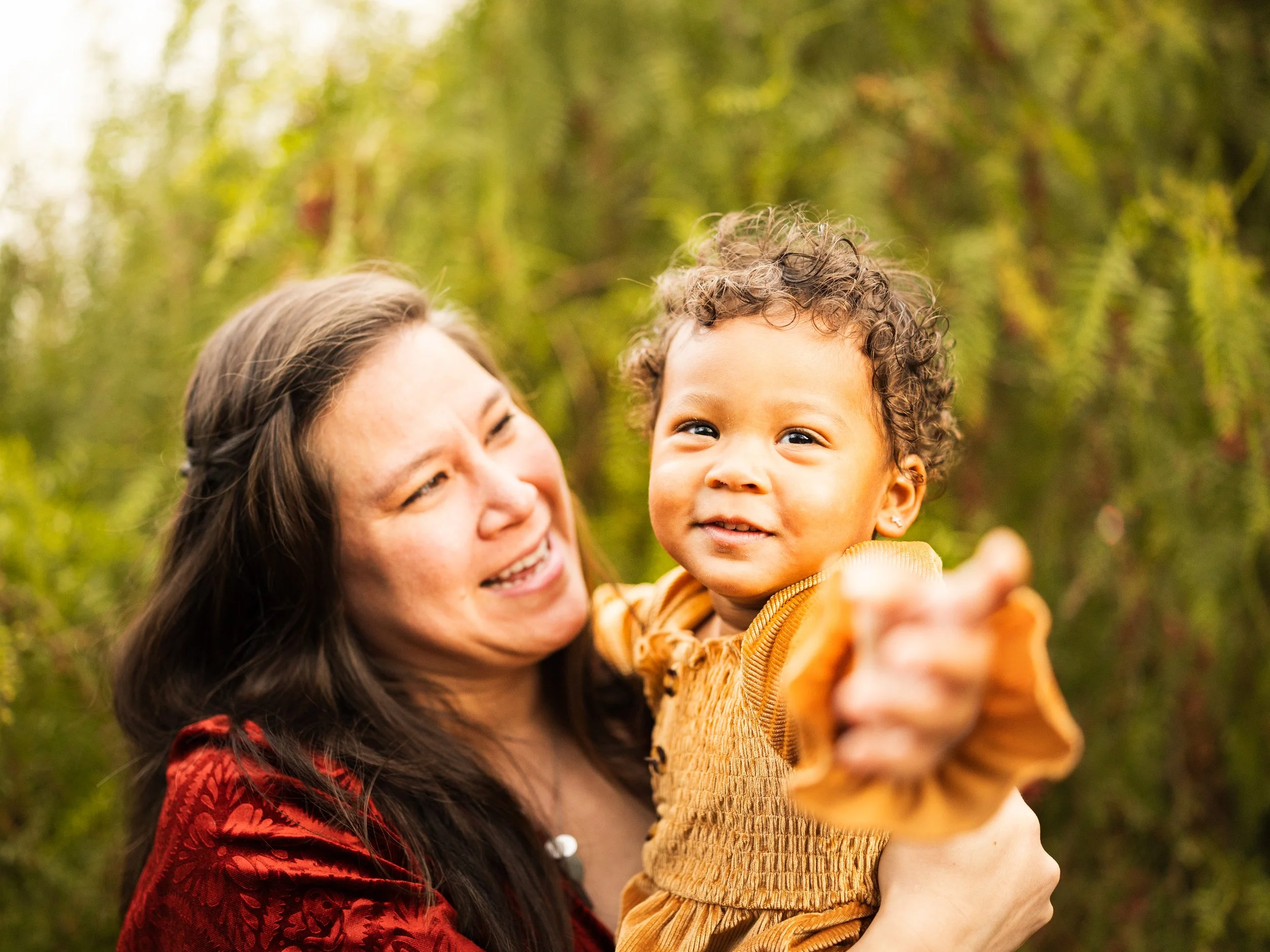 A woman holding a young girl outdoors with greenery in the background.