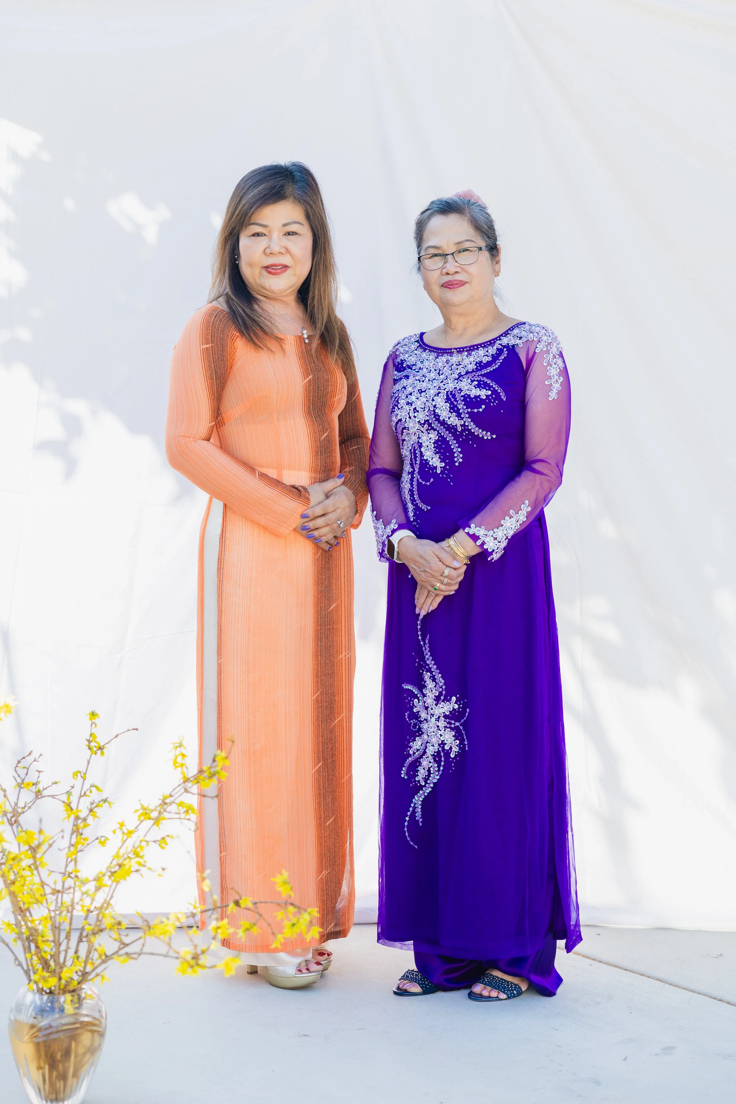 Two women standing together, one in an orange traditional dress and the other in a purple dress with embroidery, outdoors near a white backdrop.