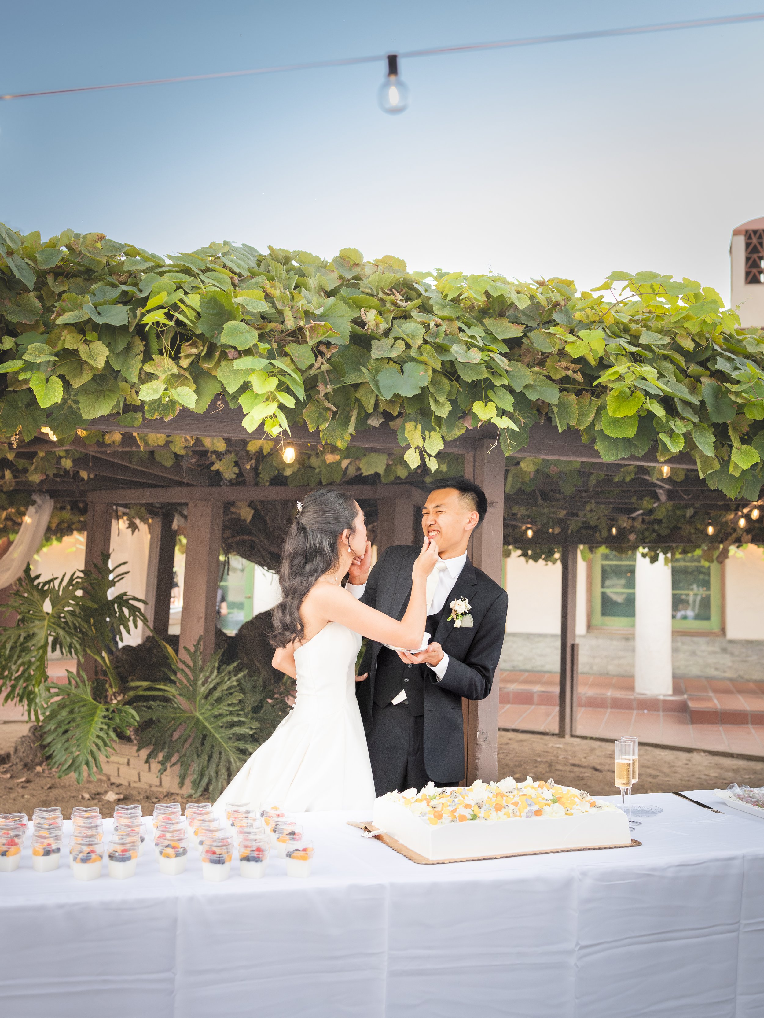 A bride and groom smiling and sharing a playful moment during their wedding reception under a vine-covered pergola. The table in front of them has a wedding cake and dessert cups.