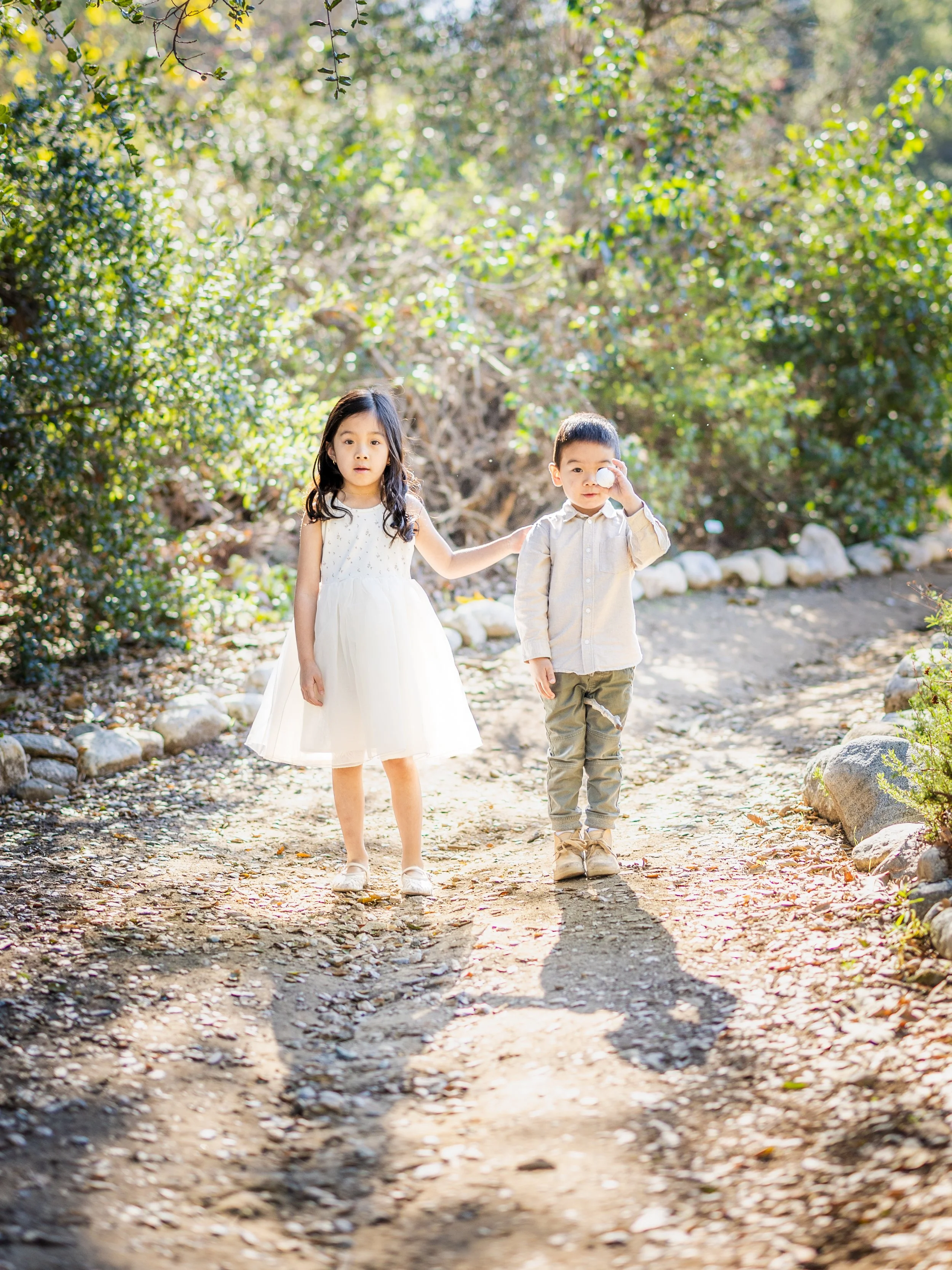 Two young children, a girl in a white dress and a boy in a beige shirt and pants, walking on a dirt path surrounded by greenery. The girl has her hand on the boy's shoulder, and the boy is holding a small ball close to his eye. They appear to be in a