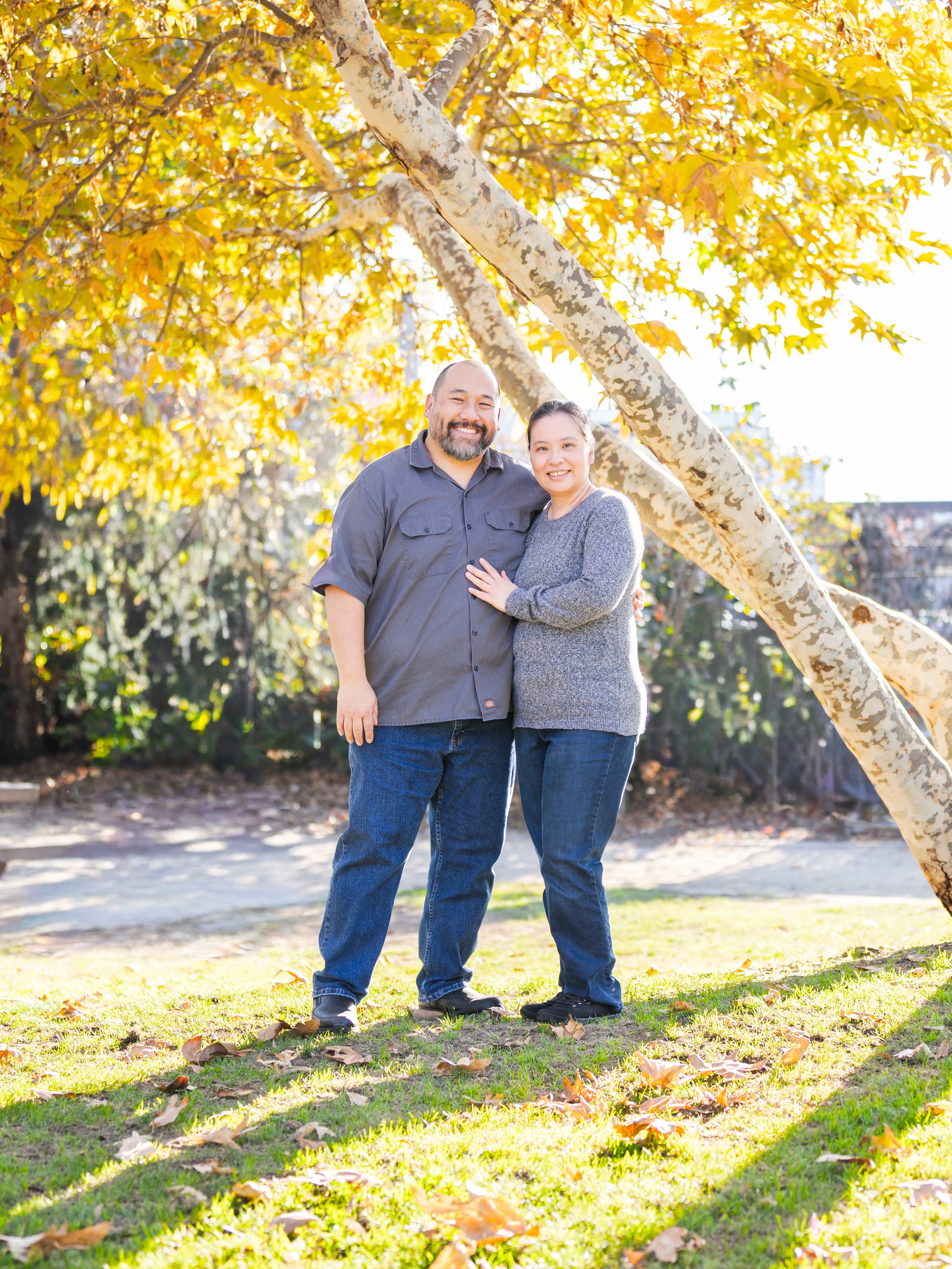 A smiling couple standing outdoors under a large, leaning tree with yellow leaves, in a park-like setting during autumn.