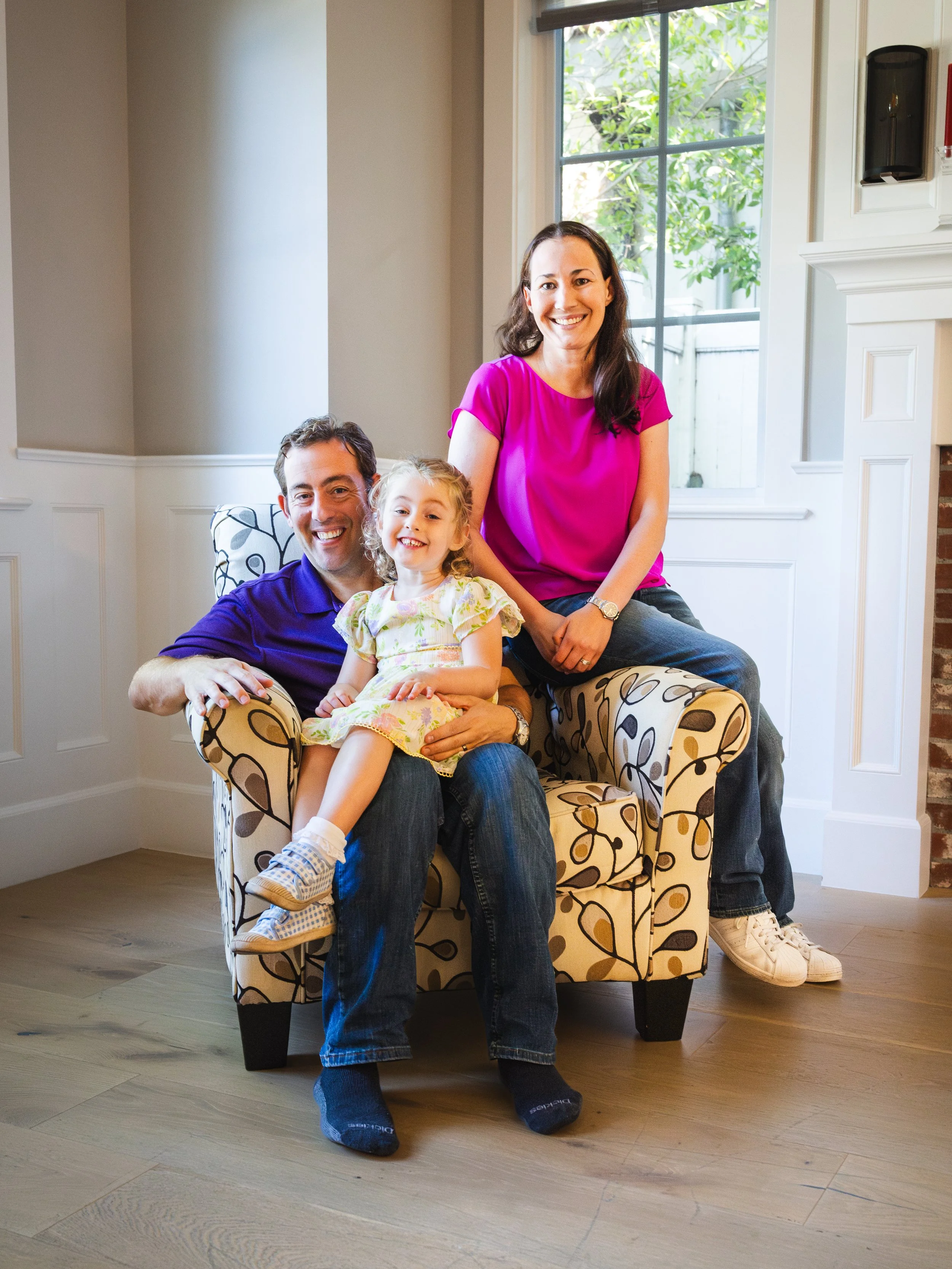 Happy family of three with a man, woman, and young girl posing together on a patterned chair in a bright room with a window and greenery outside.