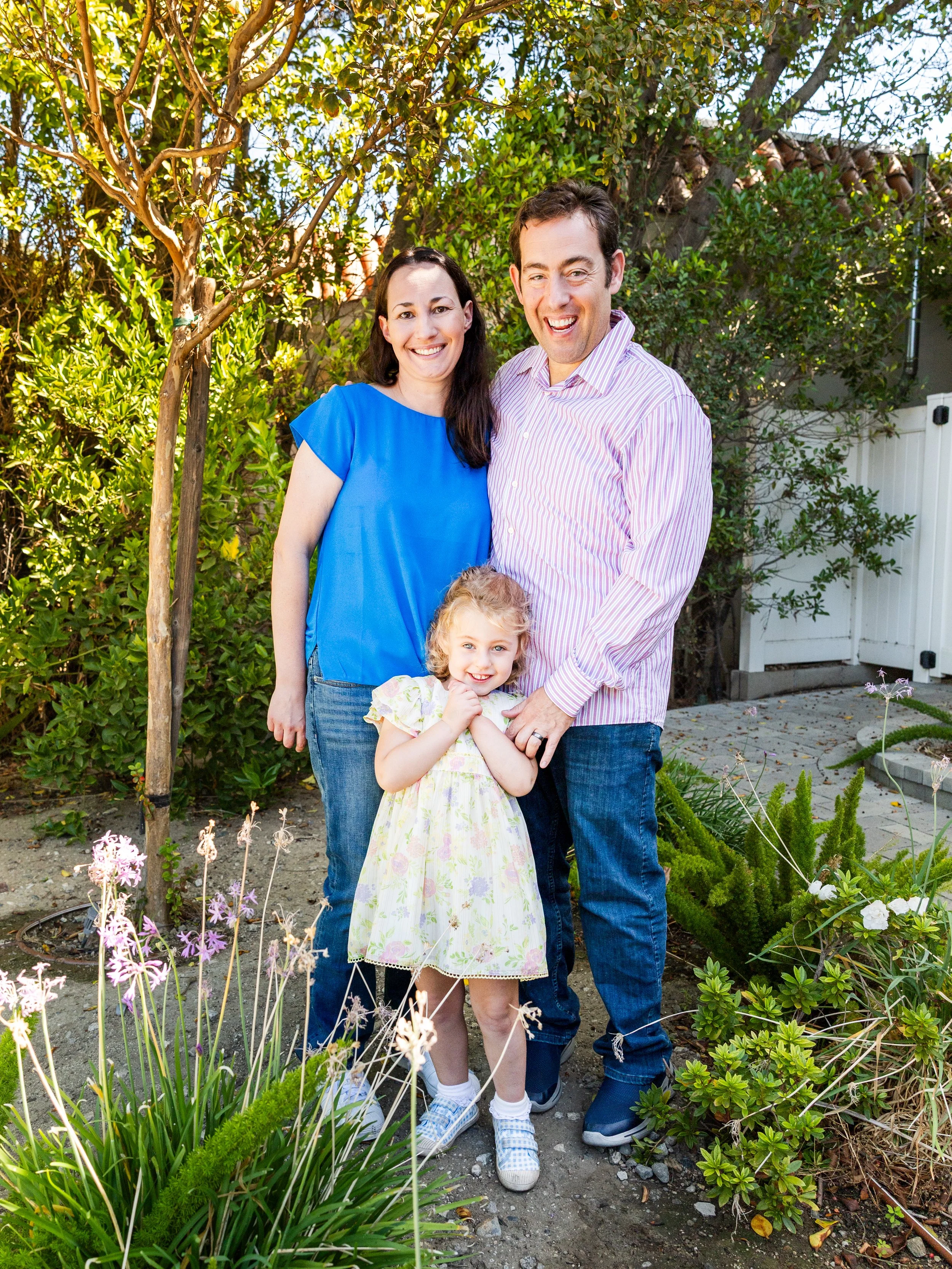 Family of three standing outdoors in a garden, smiling at the camera. The mother is in a blue top, the father in a striped shirt, and the young girl in a floral dress.