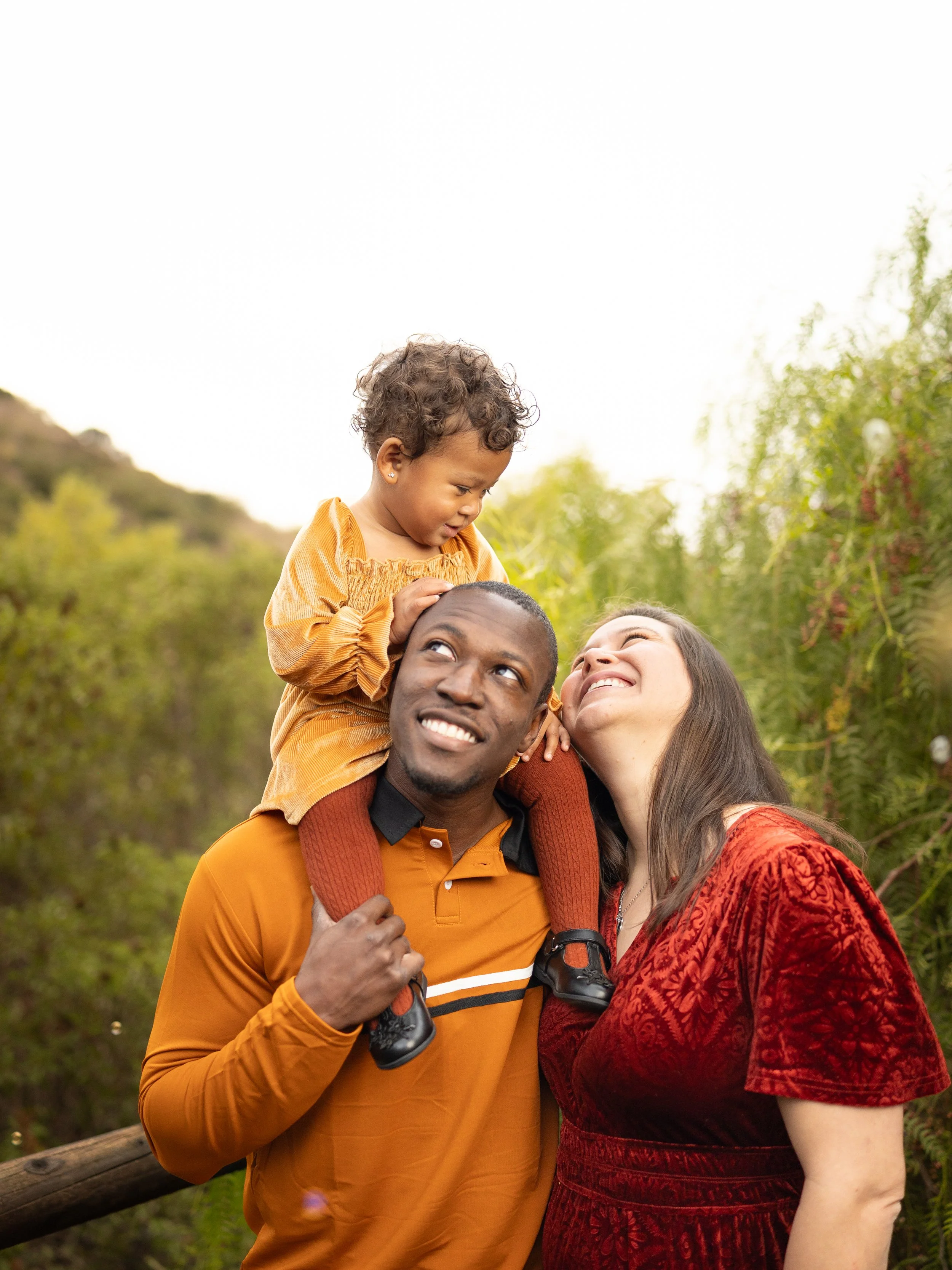 A family of three outdoors, with the man carrying a toddler on his shoulders and the woman smiling, surrounded by green foliage.