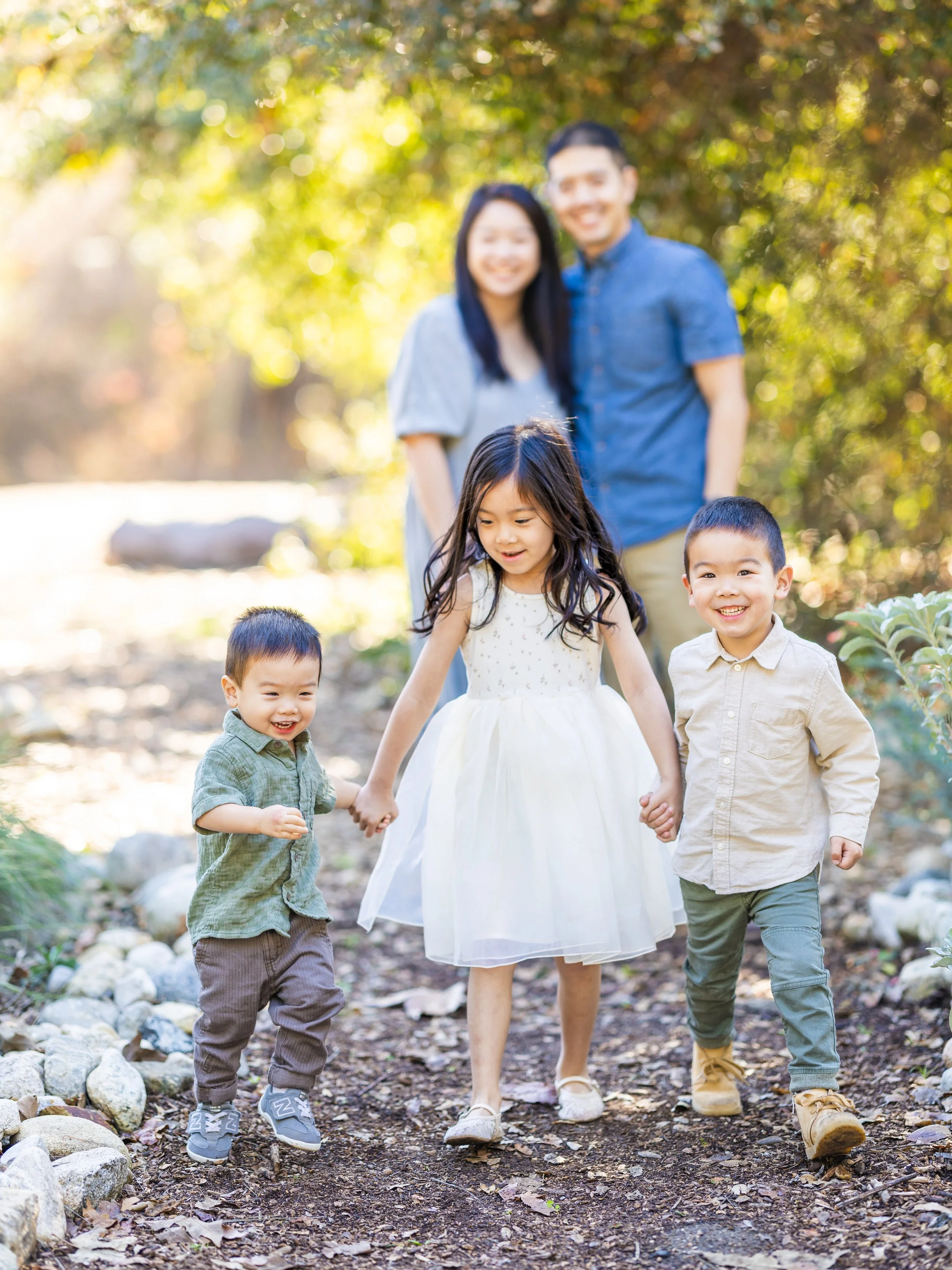 A family of five running and walking outdoors on a sunny day in a wooded area, holding hands and smiling.