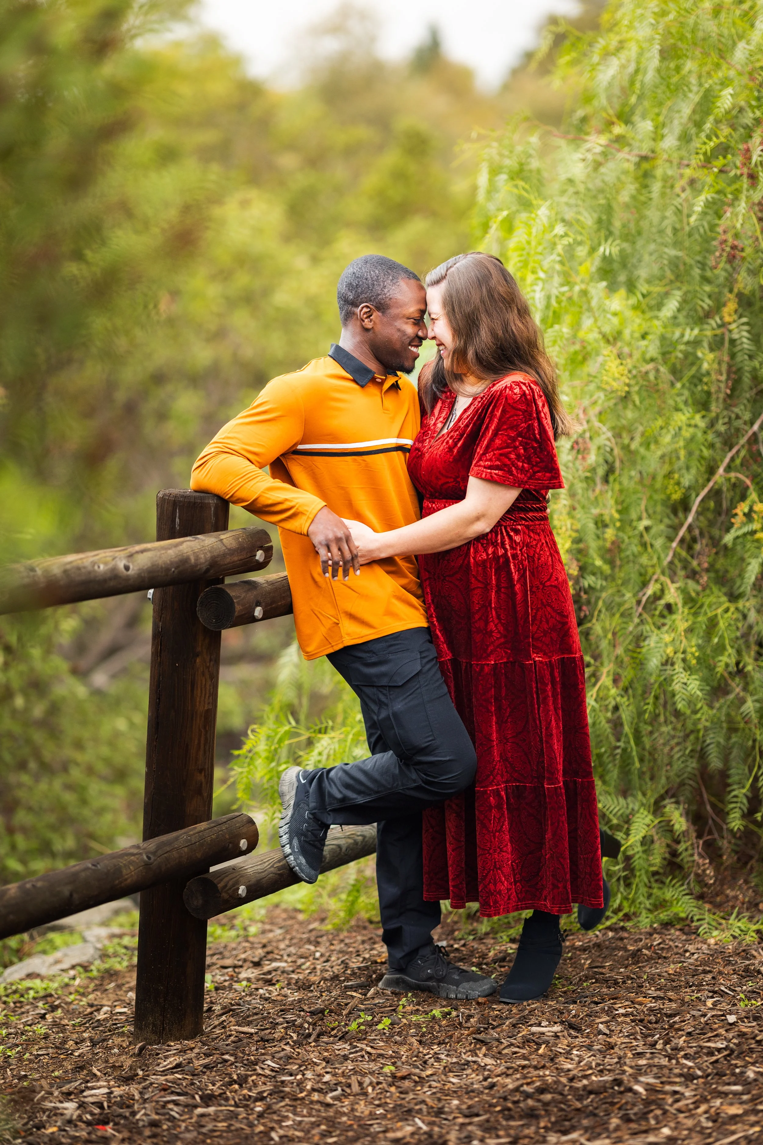 A couple sharing an intimate moment outdoors, smiling and leaning their foreheads together near a wooden fence, surrounded by green foliage.