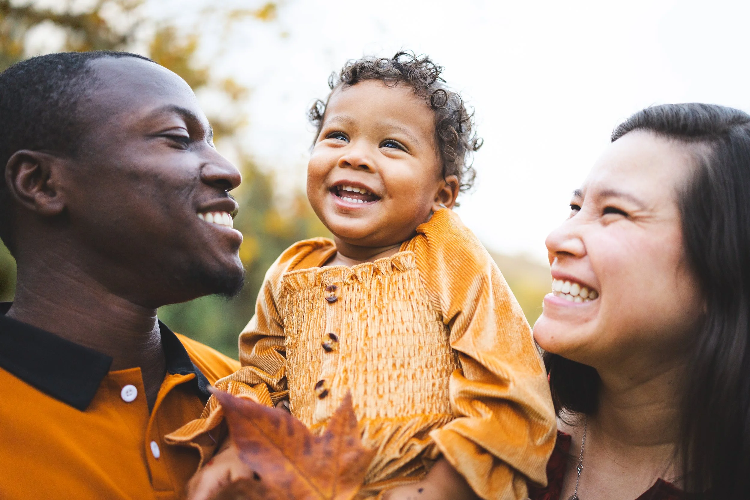 A happy family outdoors with a smiling African American man, Asian woman, and a young child with curly hair, wearing an orange outfit, holding autumn leaves.