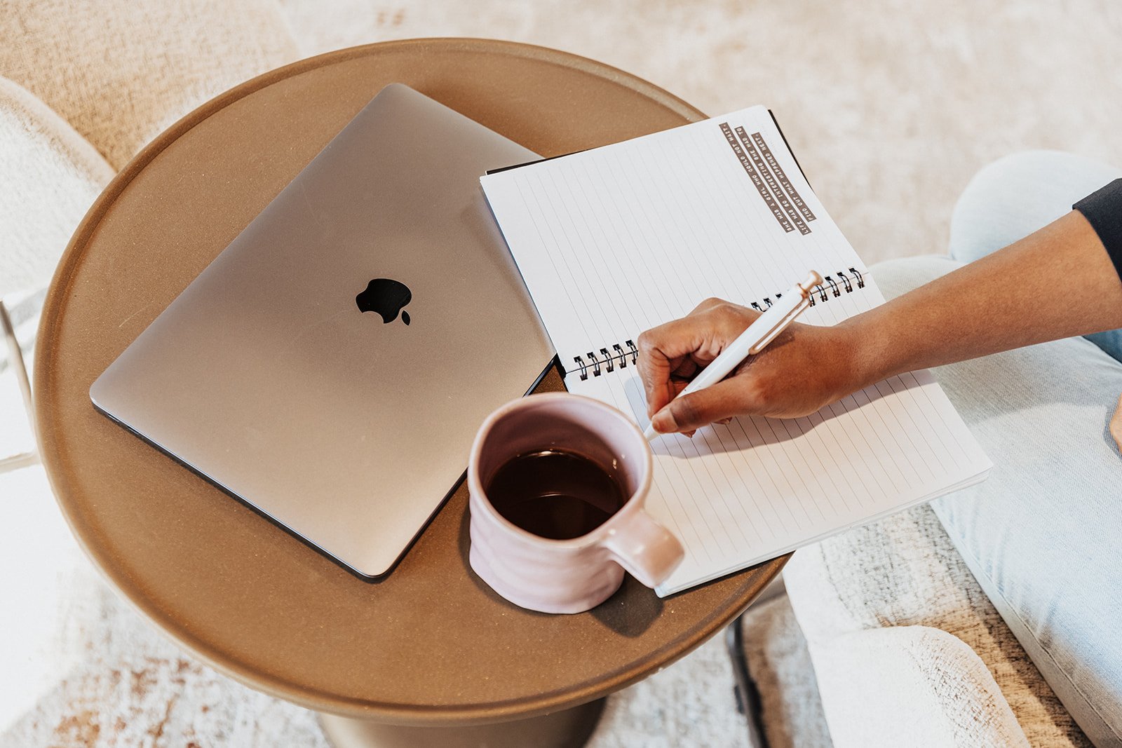 A laptop closed on a small round table next to a pink mug filled with coffee, with a person writing in a notebook with a pen, in a cozy indoor setting.