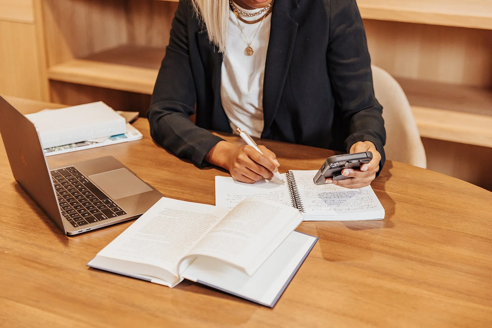 A woman sitting at a wooden desk with an open laptop, open books, and notebooks, writing in a notebook with a pen while looking at her phone.