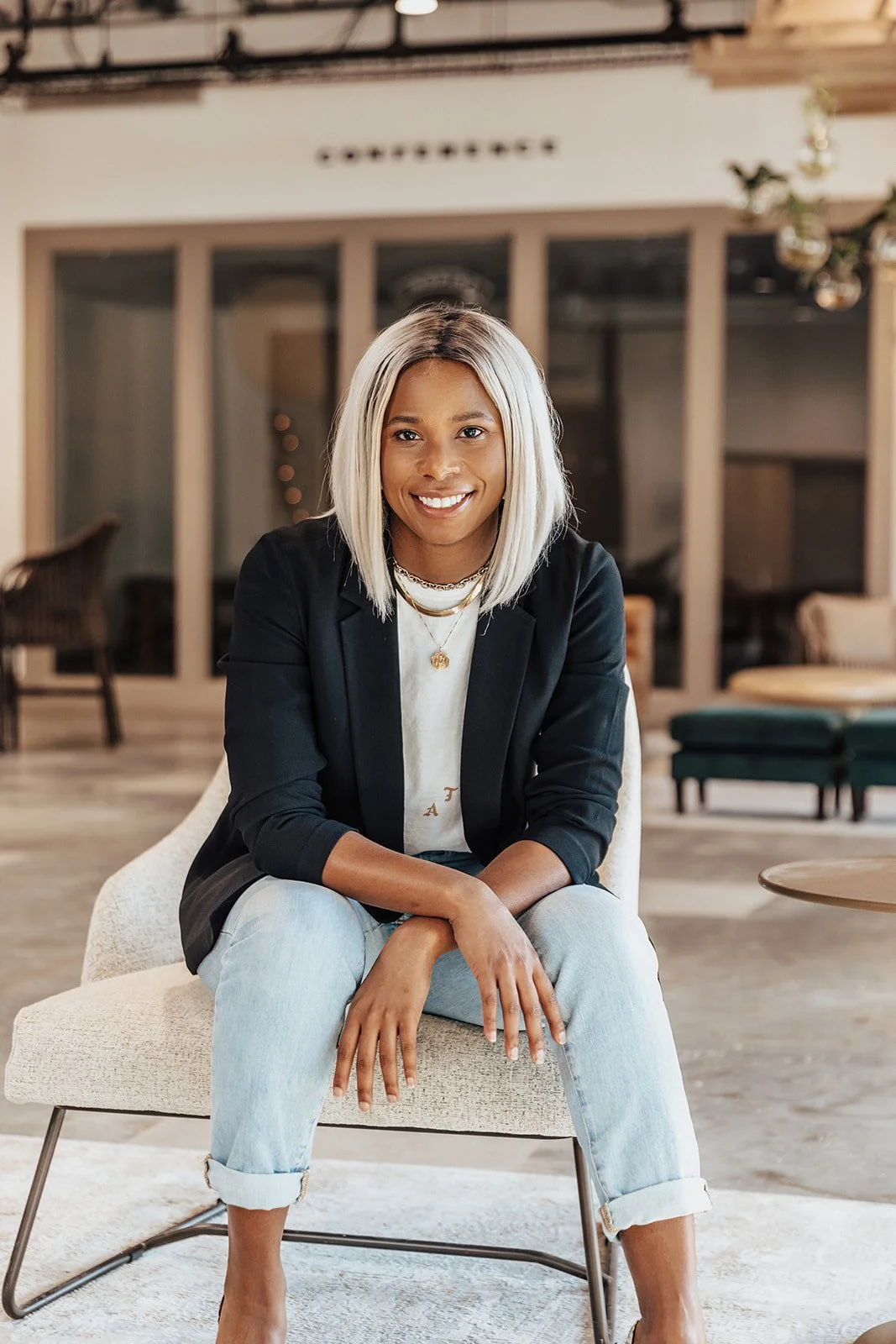 Young woman with blonde hair sitting on a light-colored chair, smiling, in a modern indoor setting with glass doors and furniture in the background.