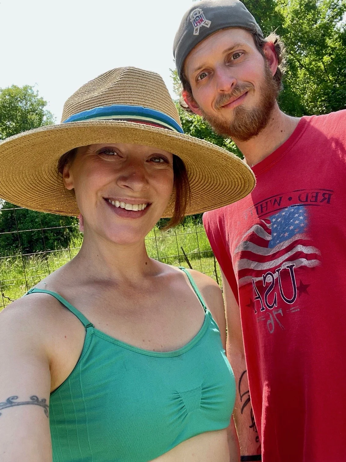 A smiling woman wearing a wide-brimmed straw hat and a green tank top, standing outdoors with a man wearing a grey baseball cap and a red T-shirt. They are in a grassy area with trees behind them on a sunny day.