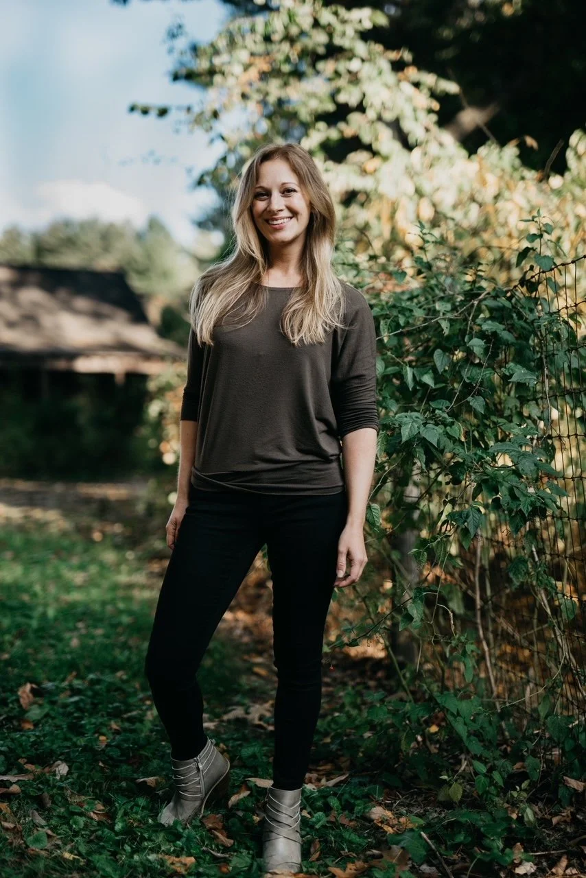 A young woman with long blonde hair smiling outdoors, standing on grass near a vine-covered fence with trees and a blue sky in the background.