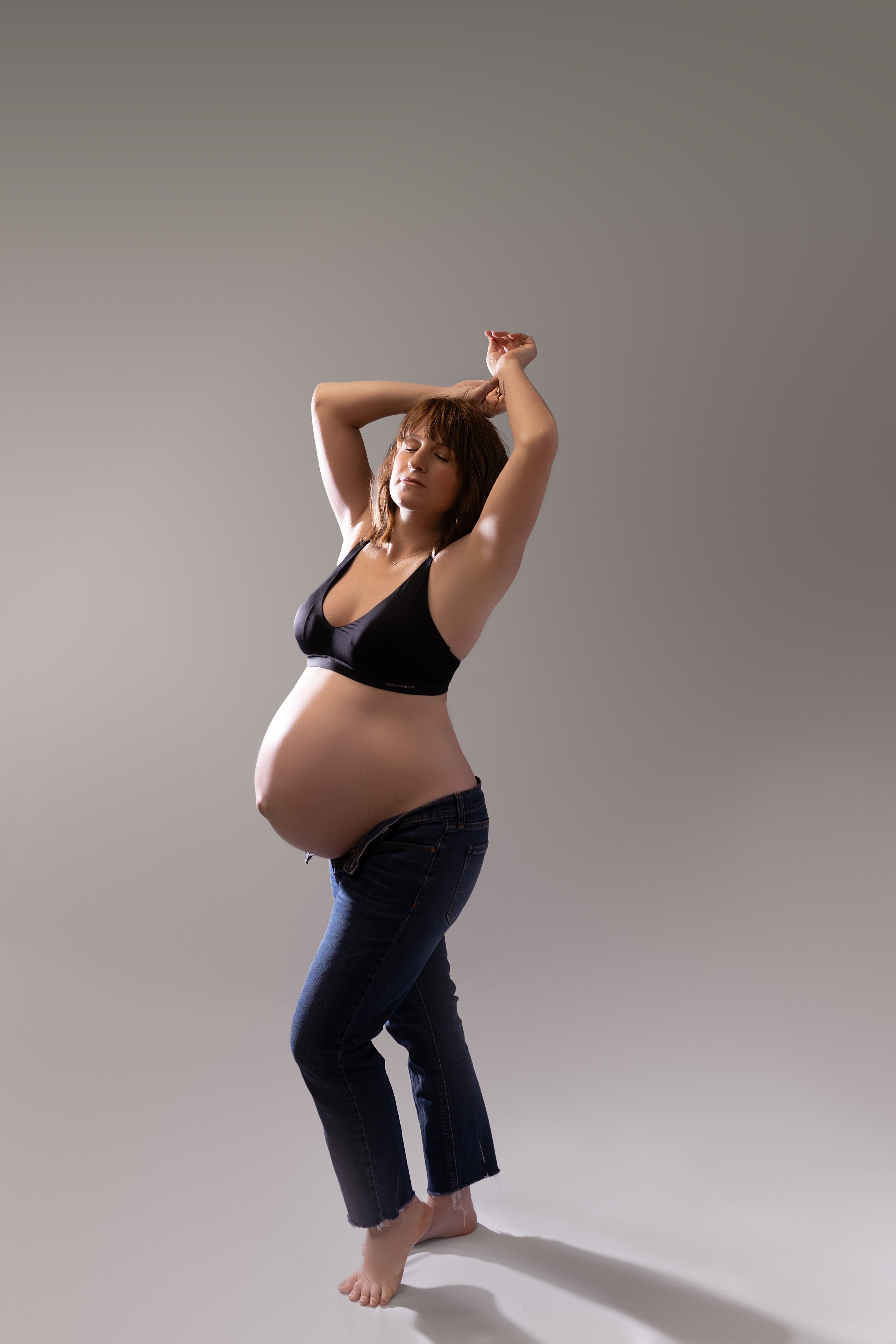 Pregnant woman with brown hair in a black sports bra and blue jeans, standing barefoot with her arms raised and eyes closed against a plain background.