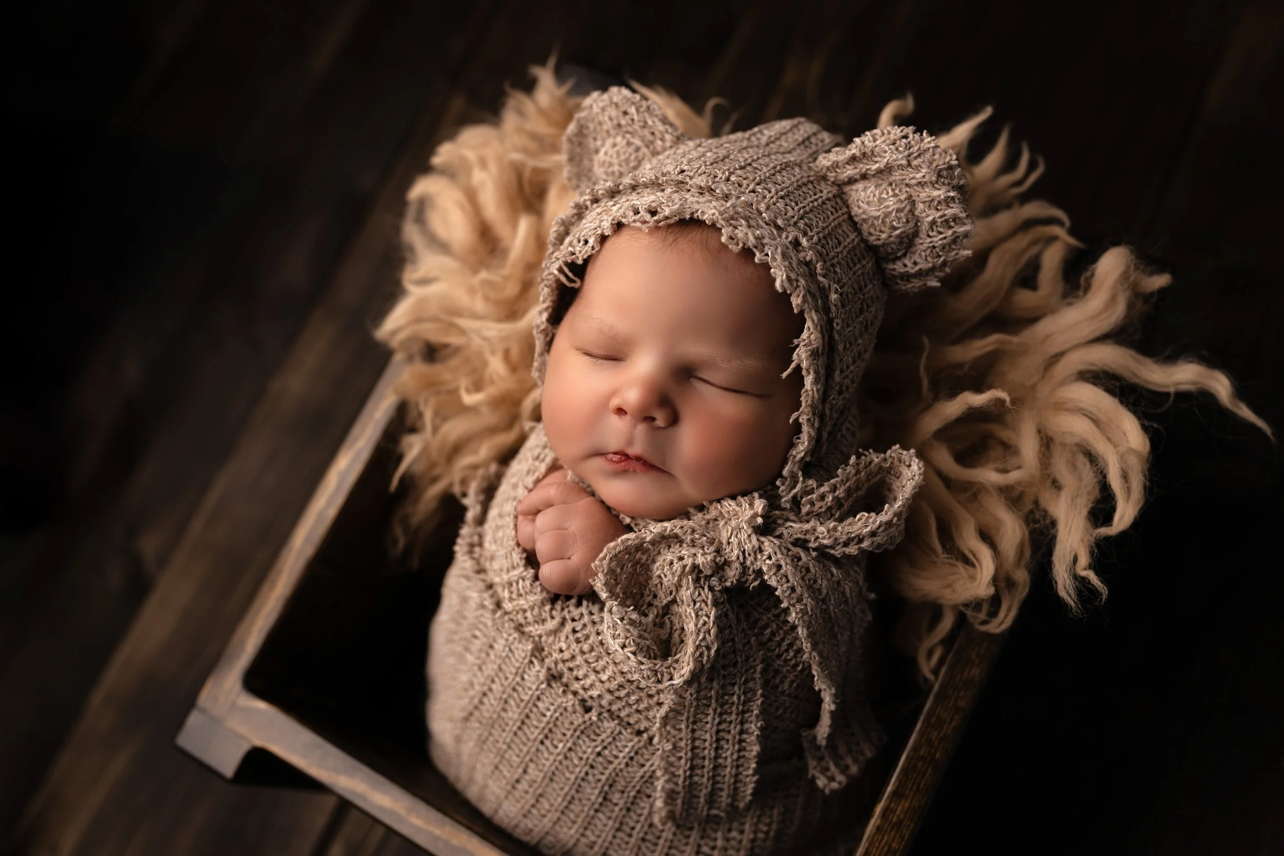 A sleeping baby wearing a knitted bear hat and sweater, lying on a fluffy rug inside a wooden box.