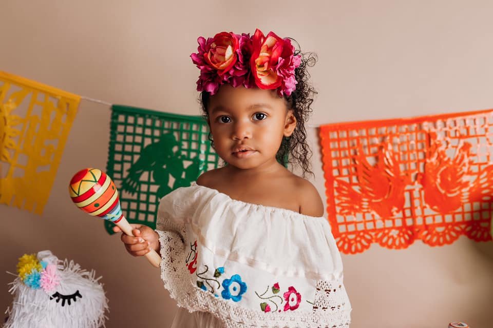 Young child wearing a floral headband and holding a maraca, standing in front of colorful papel picado decorations.
