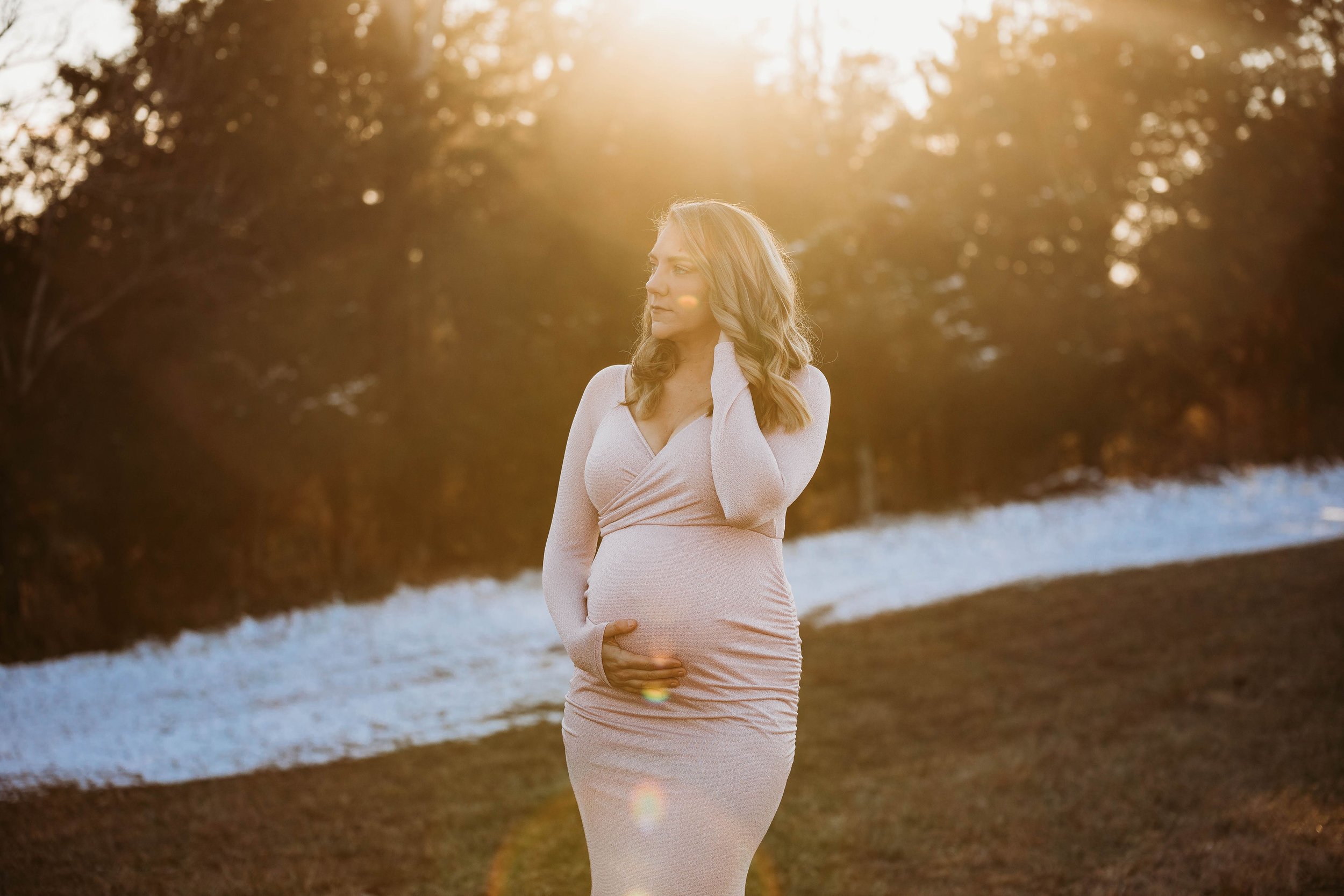 A pregnant woman in a light-colored dress standing outdoors at sunset near a river, holding her belly and touching her neck, with trees in the background. Louisville ky maternity photographer