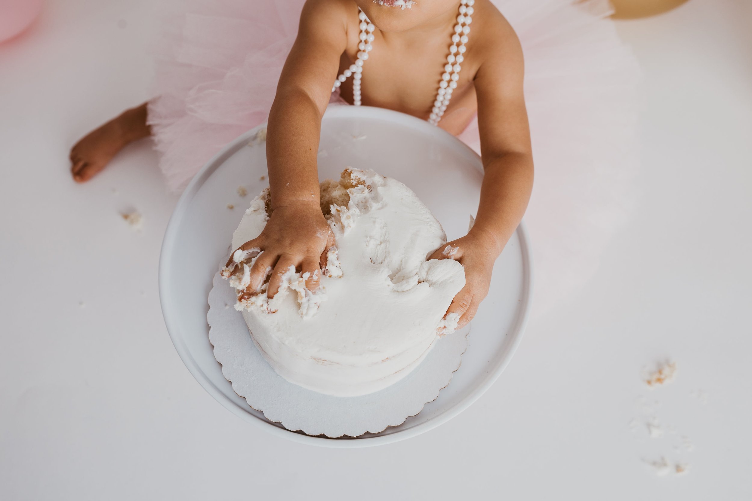 Baby wearing pink tutu, smashing hands into white frosted cake on a stand.