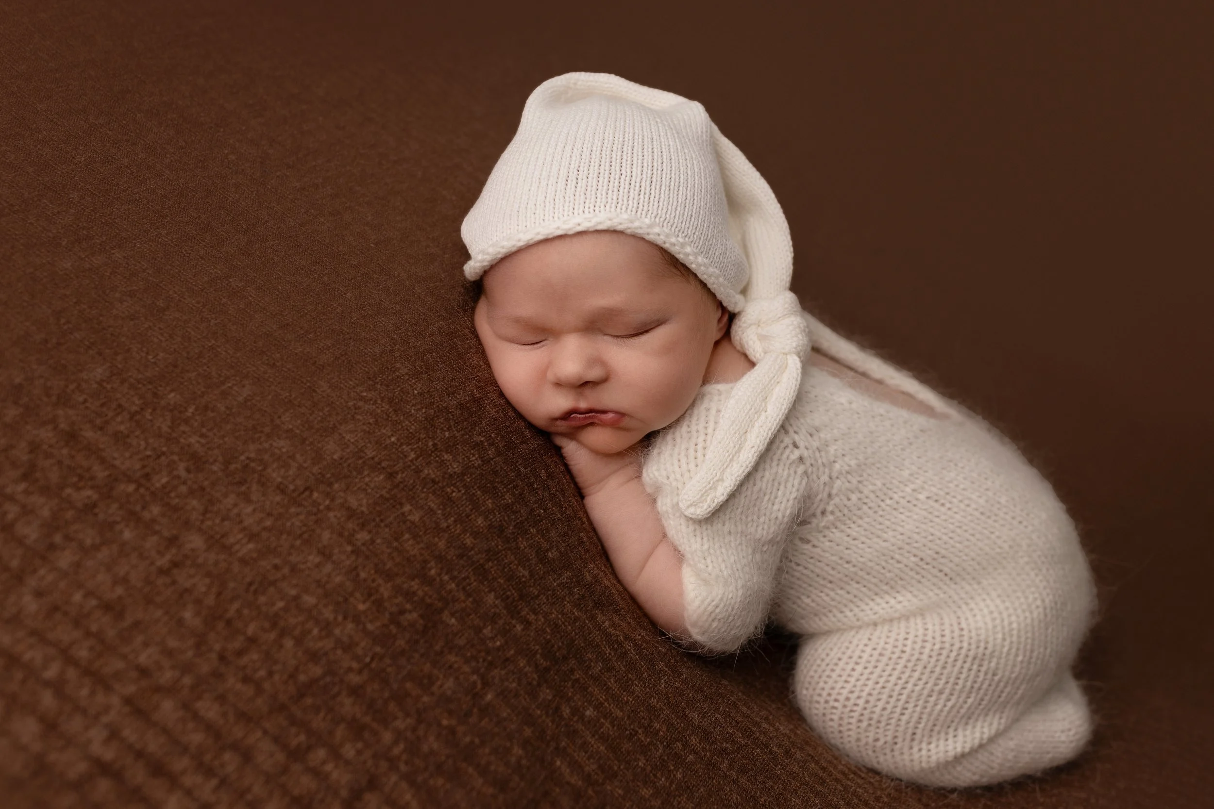 Adorable newborn baby dressed in cream-colored knit outfit and hat, sleeping on a brown sofa with head resting on hands. Louisville Kentucky Newborn Photographer