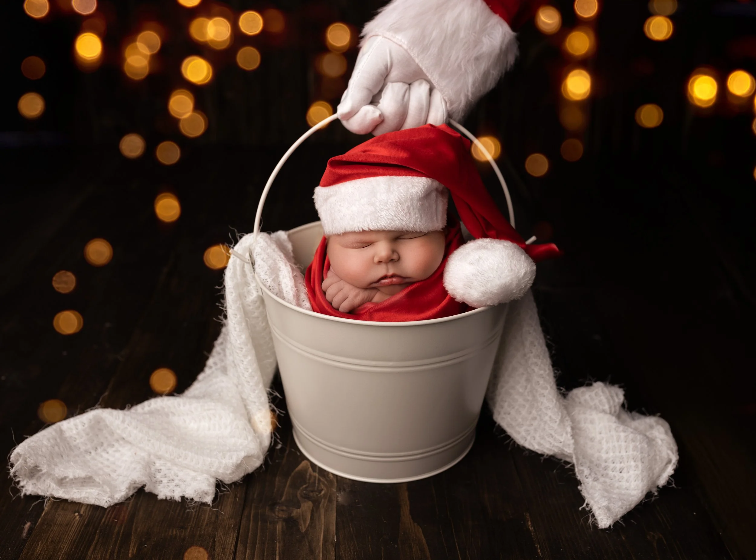 A sleeping baby dressed as Santa Claus, placed in a white bucket. The baby wears a red Santa hat with white fur trim and a red cloak. A white glove is gently holding the hat's pom-pom above the head. The background has warm, blurry string lights, cre