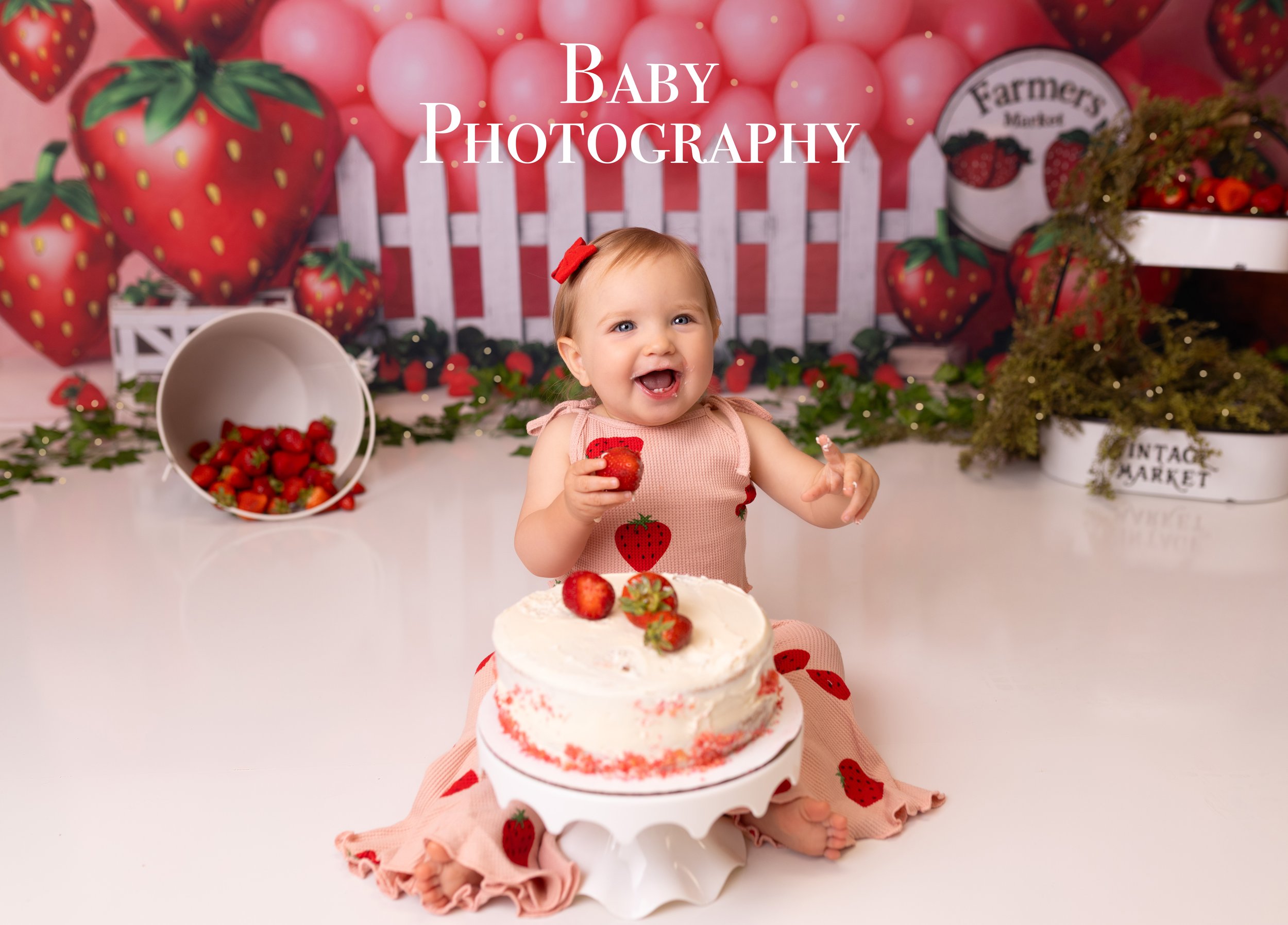 A smiling baby girl sitting in front of a strawberry cake, holding a strawberry, with a strawberry-themed backdrop and strawberries on the cake and surrounding area.Louisville Kentucky Baby Photographer