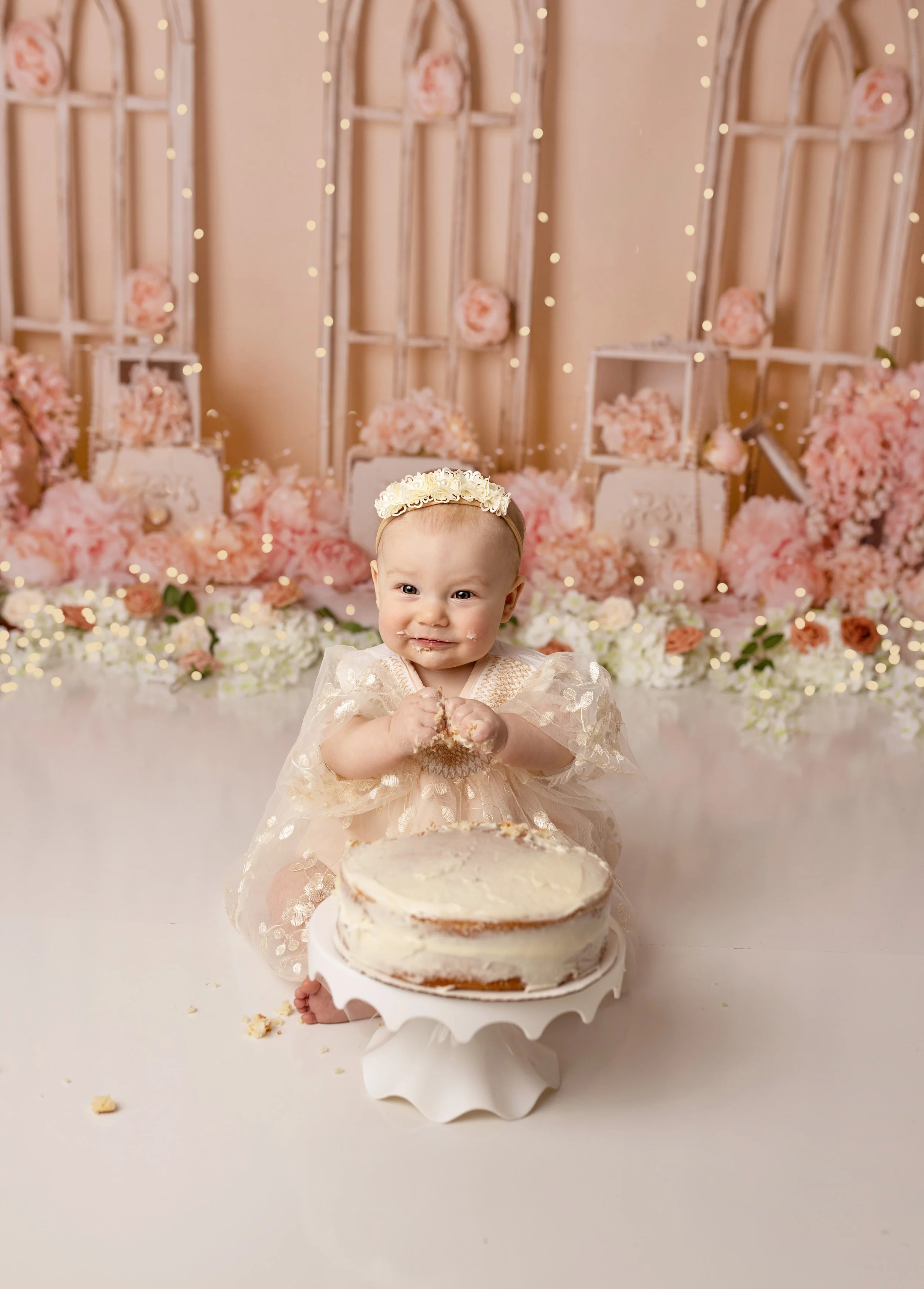 A baby girl celebrating her first birthday, sitting in front of a partially eaten cake, with a festive pink and white floral backdrop.