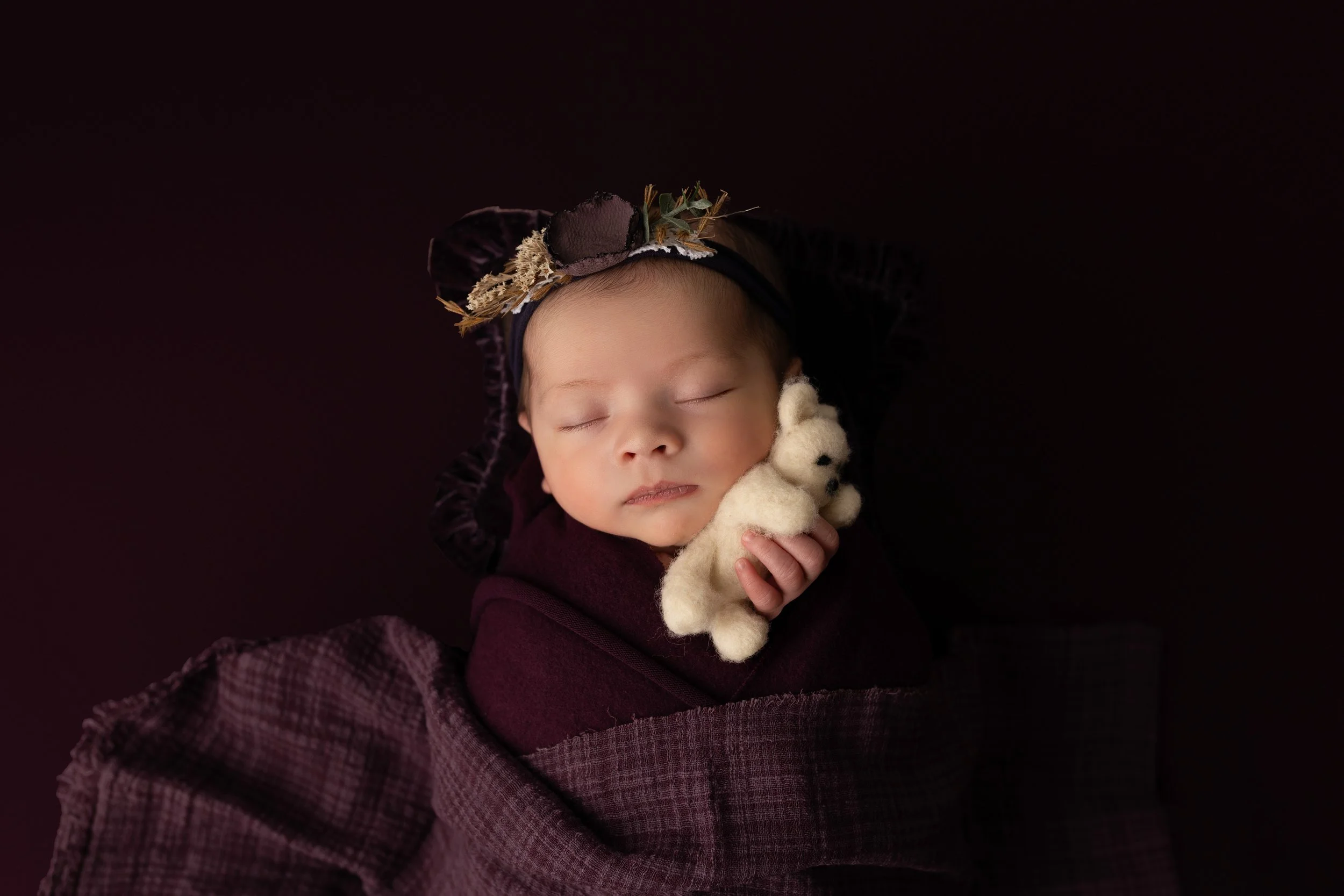 A sleeping baby wearing a dark headband with flowers, wrapped in a purple blanket, holding a small white teddy bear, against a dark background.