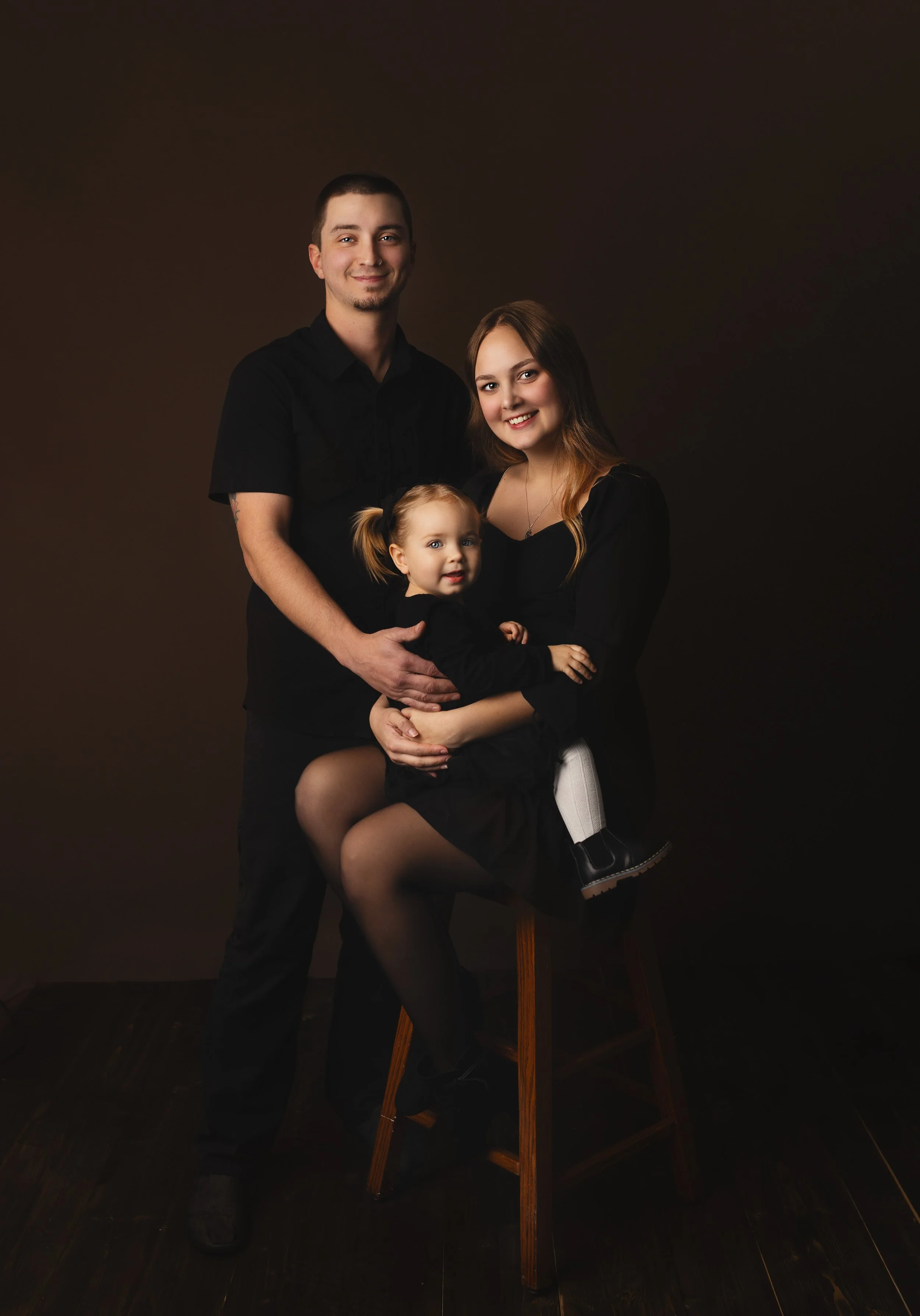 Family portrait of a man, woman, and young girl against a dark background. The man has short dark hair and is wearing a black shirt. The woman has long, wavy hair and is wearing a black dress. The young girl, sitting on the woman's lap, has blonde ha