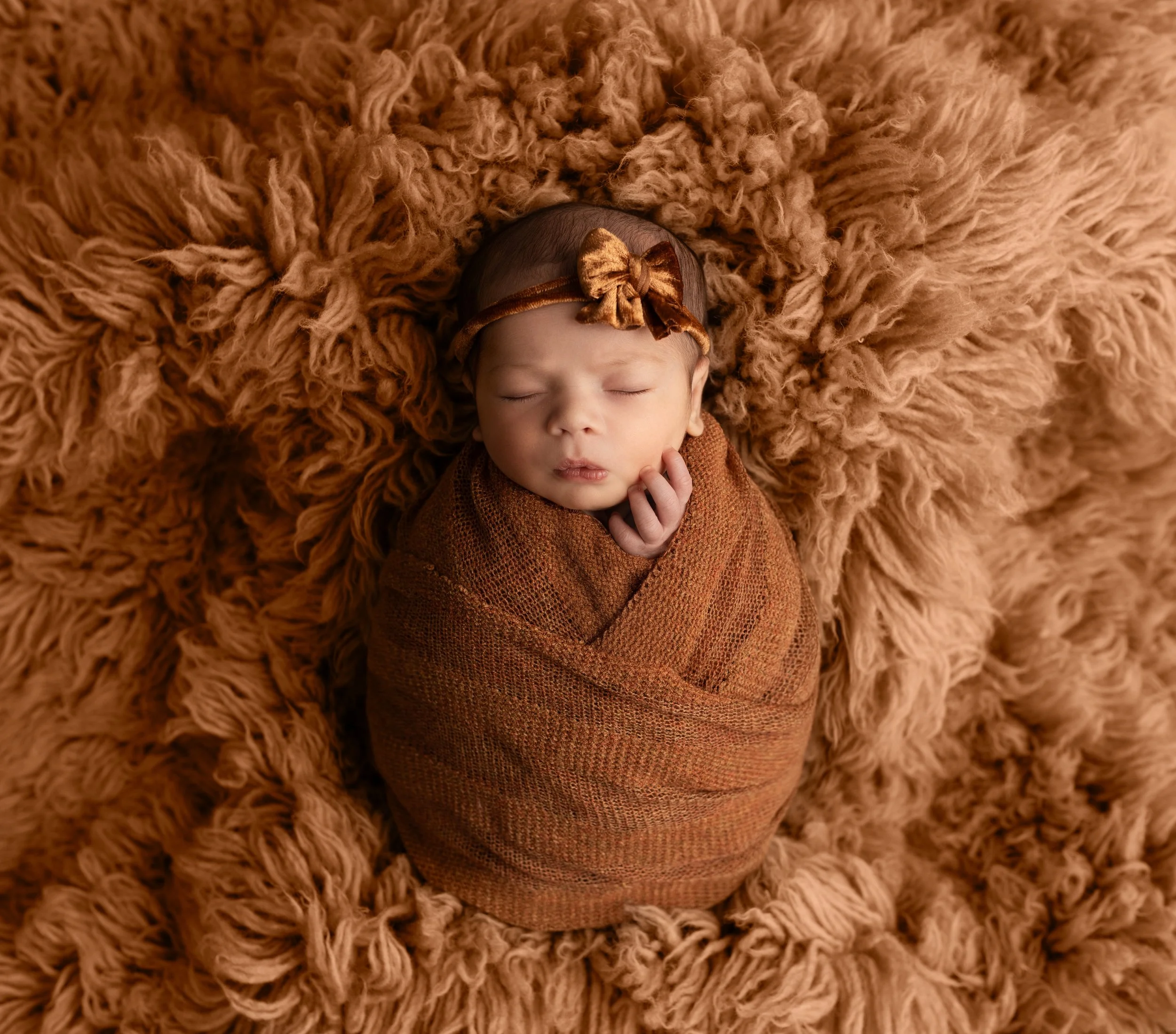 A newborn baby wrapped in a brown swaddle blanket, sleeping peacefully on a fluffy, brown textured surface, wearing a matching brown headband with a bow.