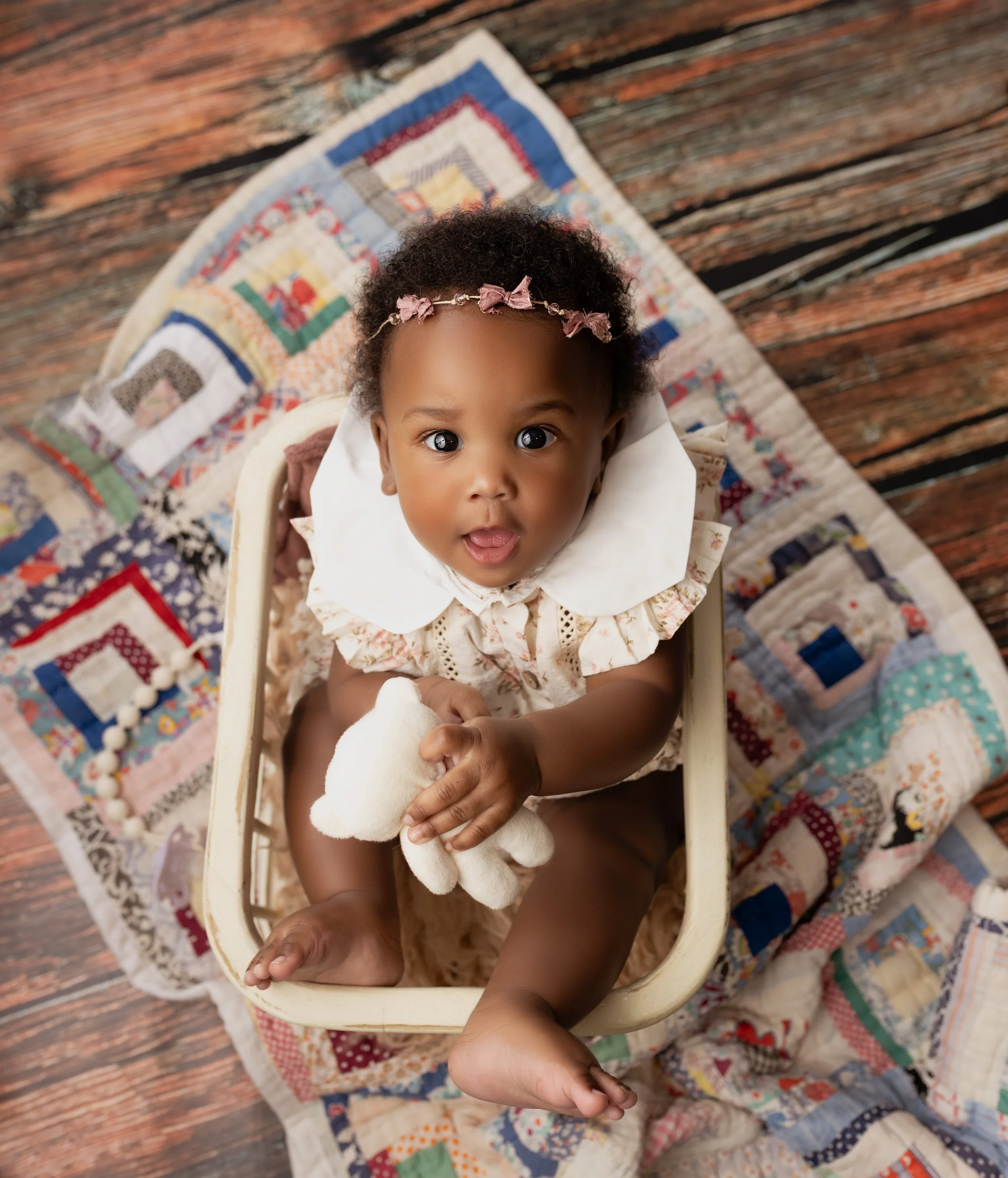 A young child with curly black hair wearing a floral dress and a pink bows headband, sitting inside a small white basket on a quilted blanket. The child holds a white teddy bear and looks up at the camera with wide eyes and an open mouth. The scene i