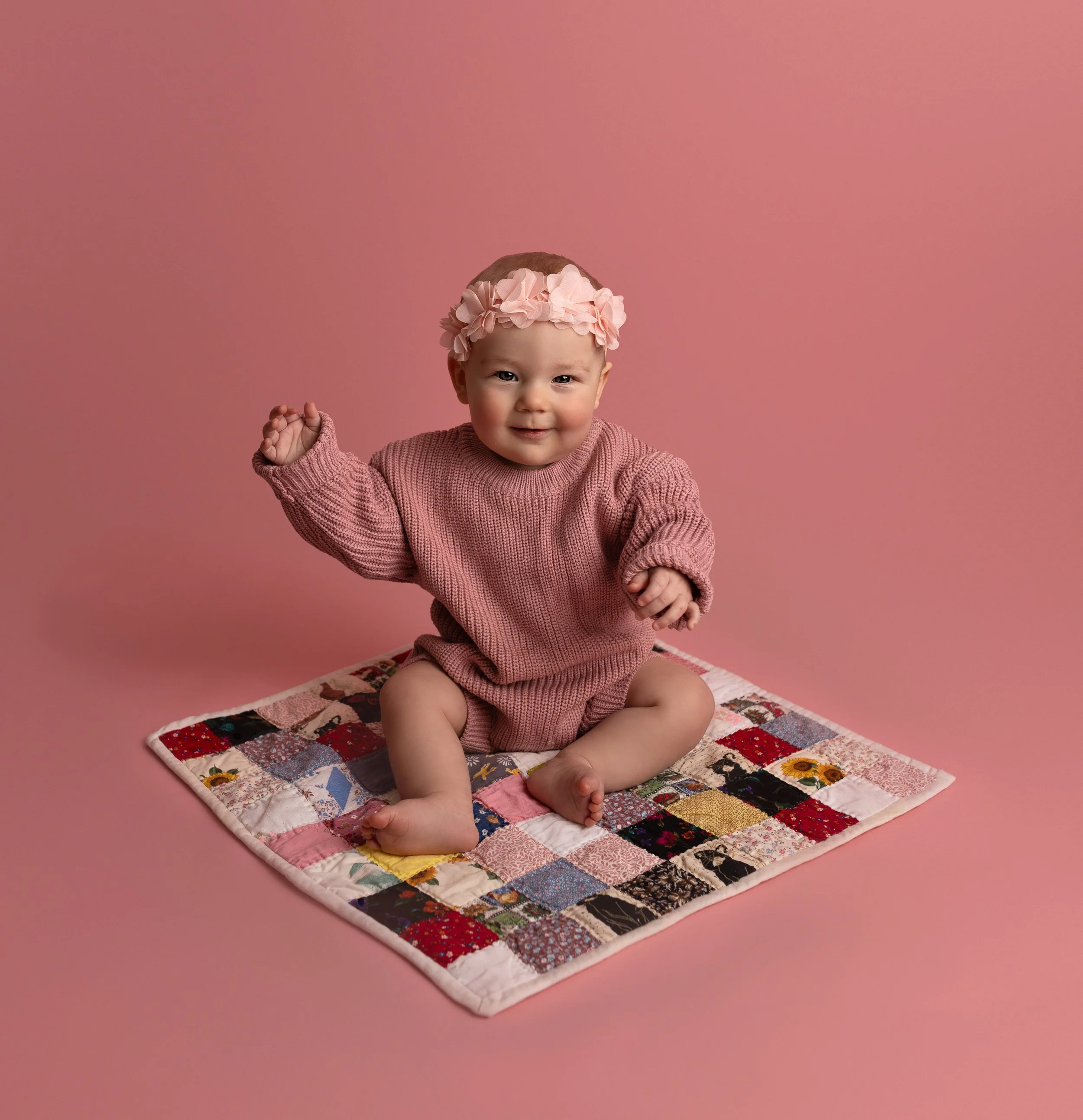 A smiling baby girl in a pink sweater, wearing a pink flower headband, sitting on a colorful quilt against a pink background.