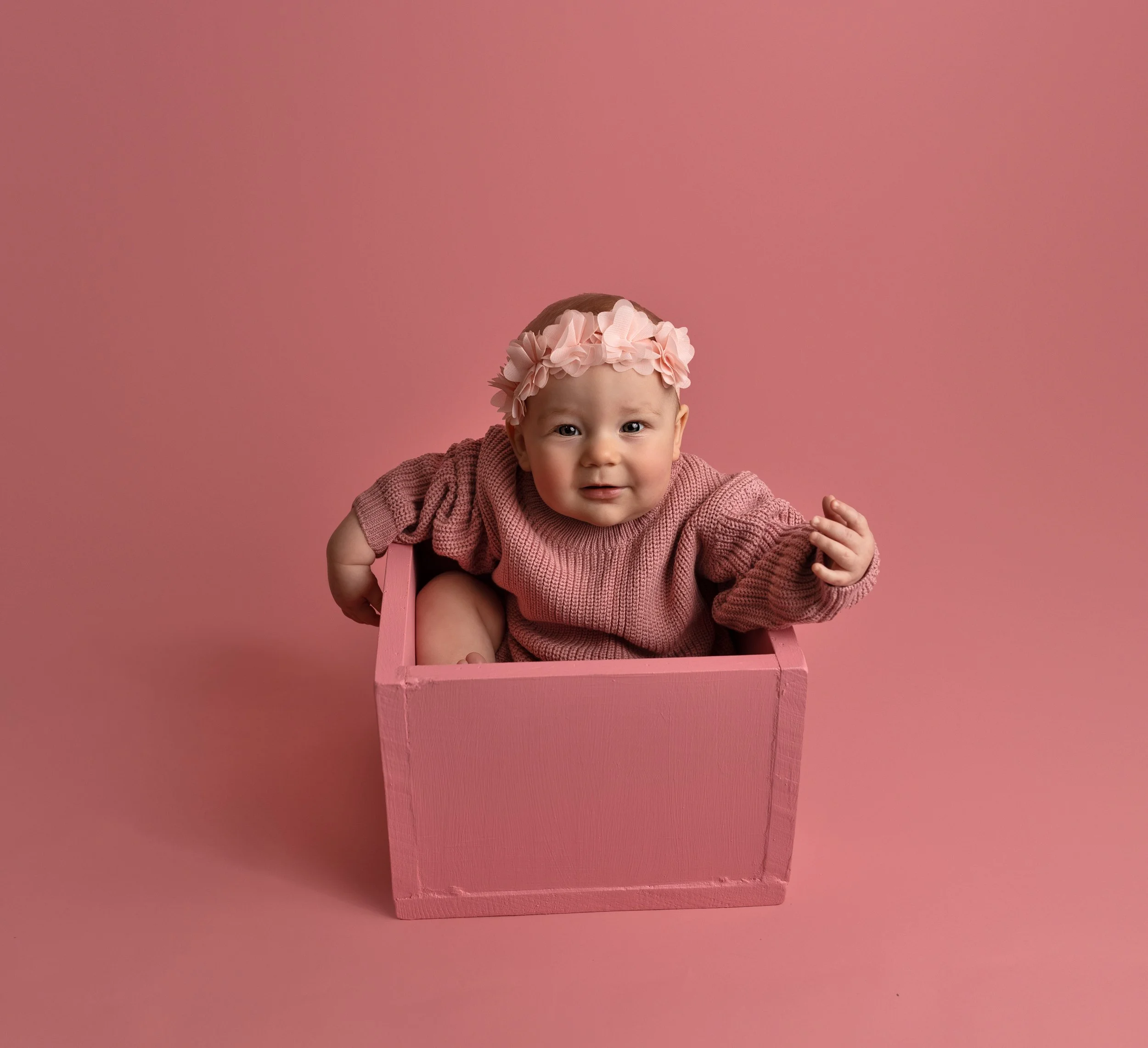 A baby girl with a pink flower headband and a knitted pink sweater sitting in a pink wooden box against a pink background.
