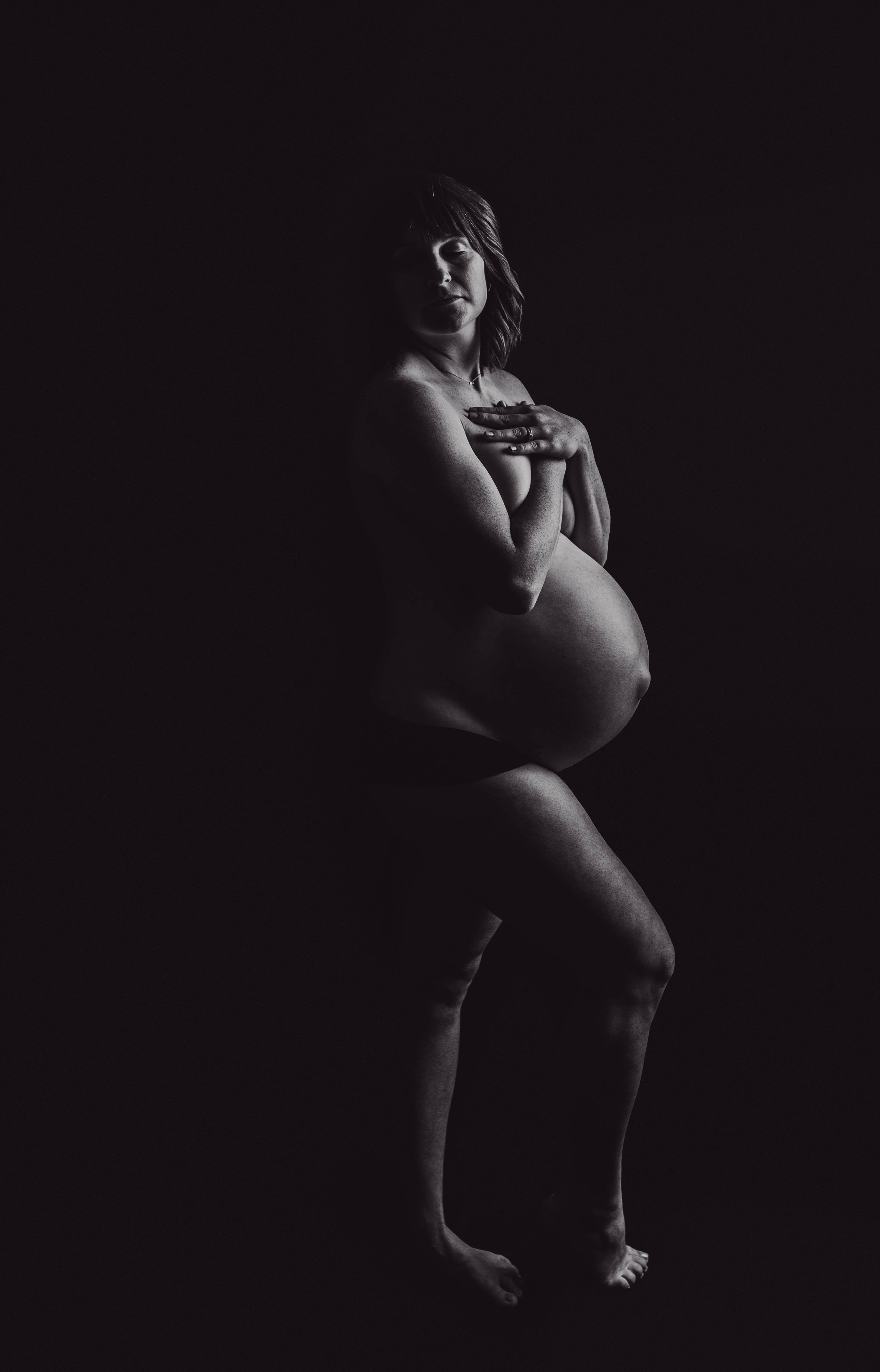 Black and white portrait of a pregnant woman, standing nude against a dark background, with one hand on her chest and the other resting on her abdomen.