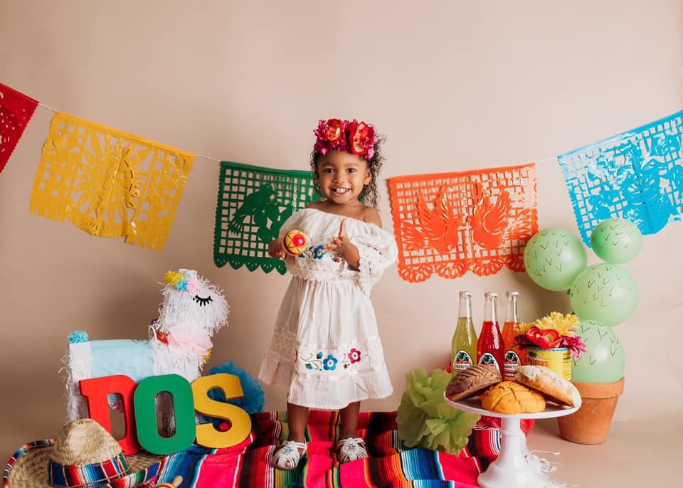 A young girl wearing a white dress and flower headband stands in front of colorful papel picado banners. She holds a maraca, with a llama piñata, sombrero, and multicolored letters spelling 'DOS' nearby. On a table, there are Mexican sodas and pastri
