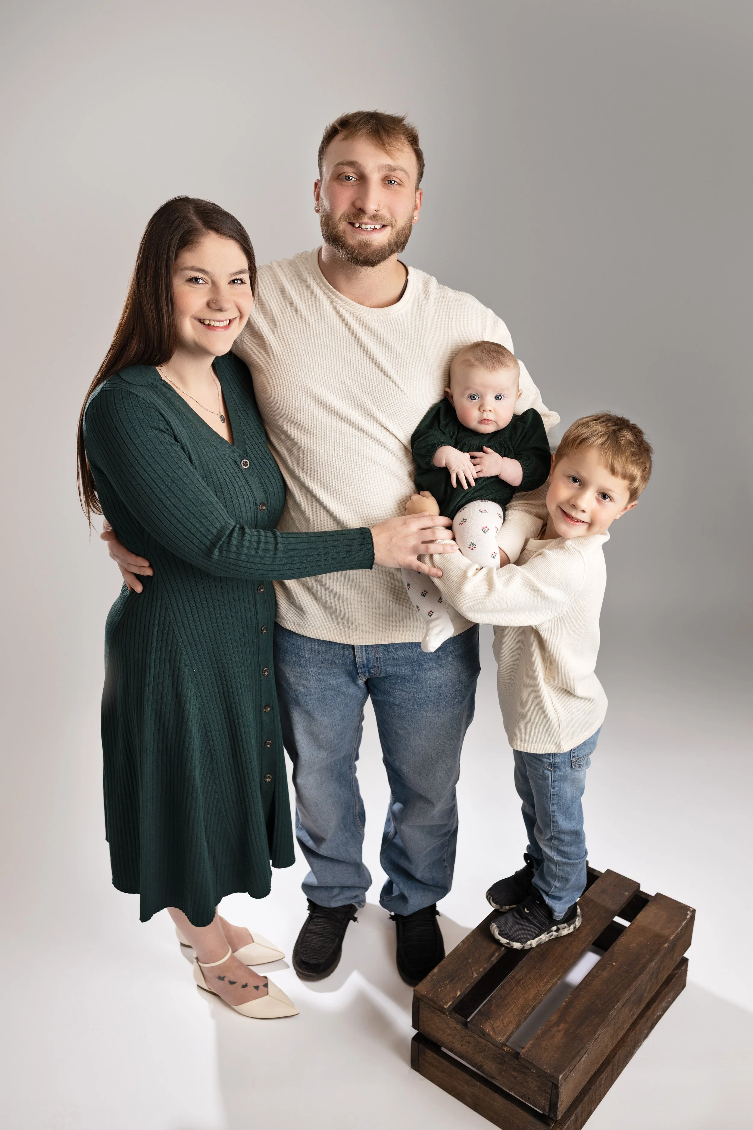 A family of four standing together in a studio with a plain background. The mother has long dark hair, wears a dark green dress, and beige shoes. The father has short hair, a beard, and wears a cream sweater with blue jeans. The daughter, a young boy