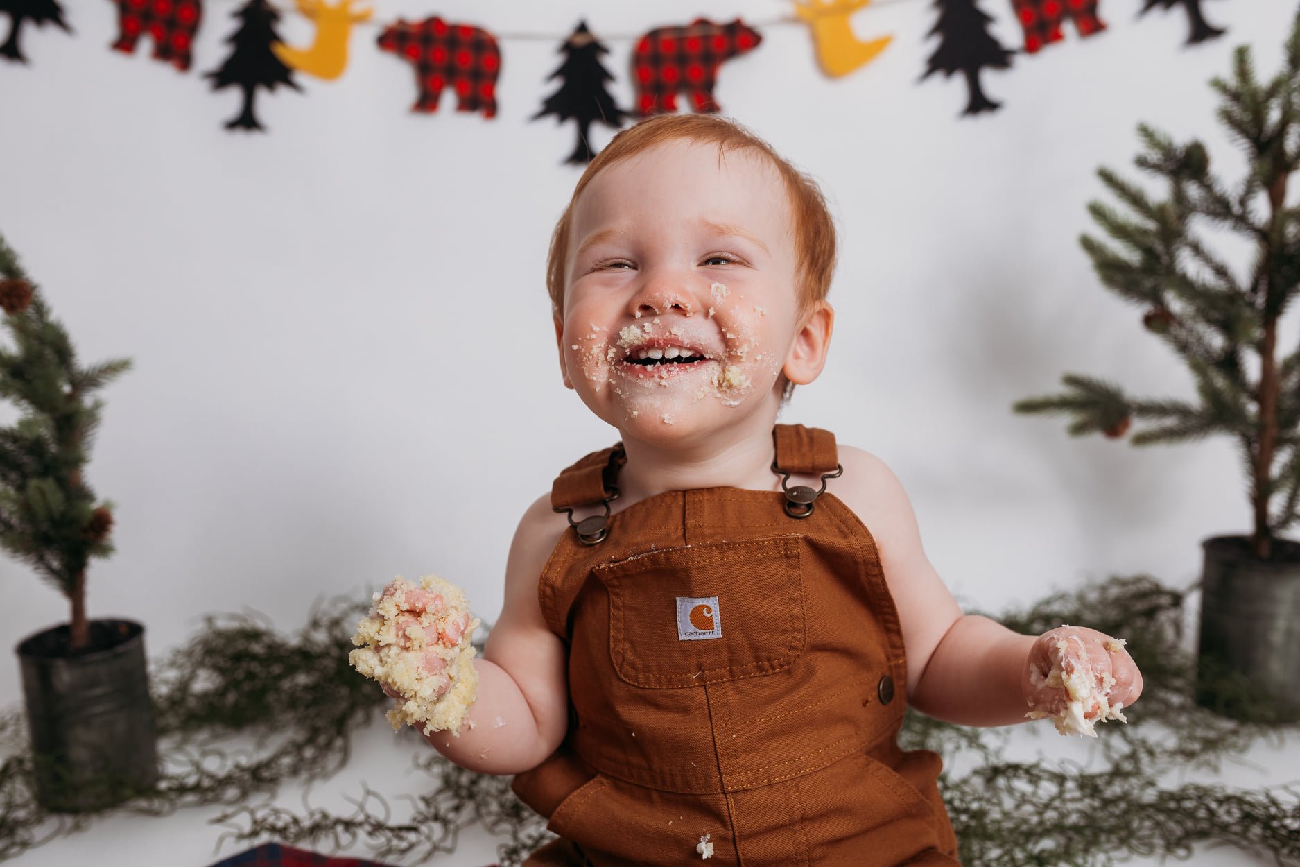 Smiling child with cake on face and hands, wearing brown overalls, sitting between small pine trees and festive banner in background.