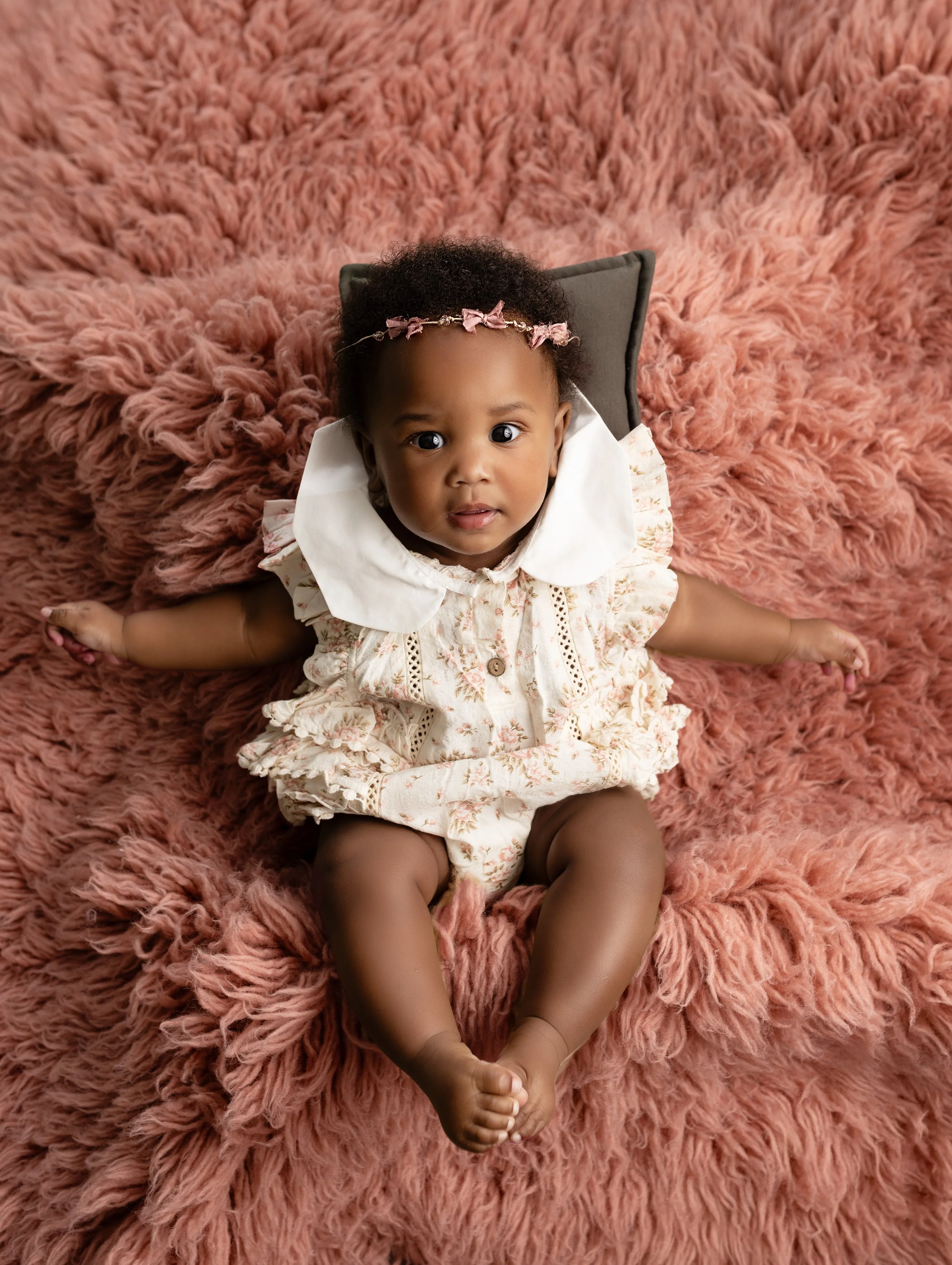 Adorable baby girl lying on a fluffy pink blanket, wearing a floral dress, with a headband with pink bows, looking up at the camera.
