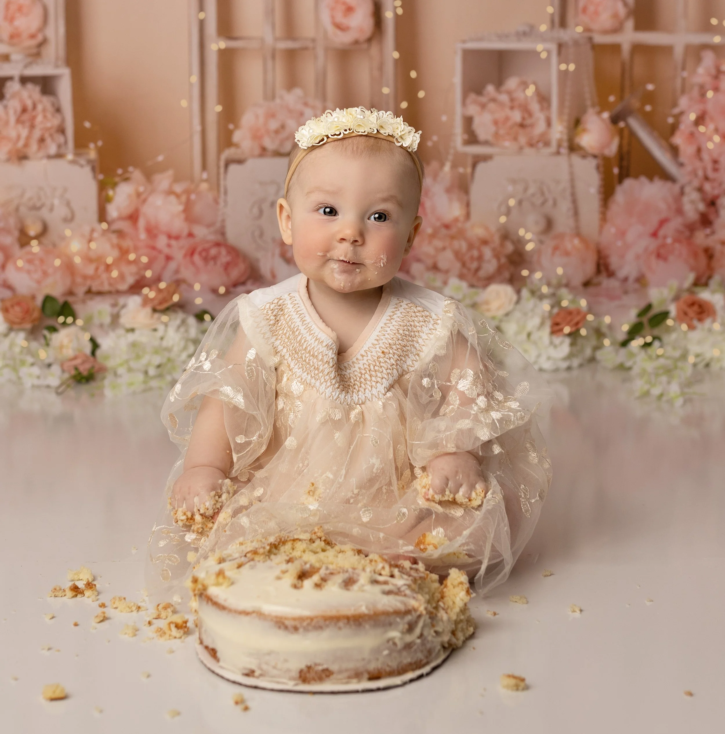 A baby girl sitting on the floor with a partially eaten cake in front of her, wearing a fancy cream-colored dress and a floral headband, in a decorated space with pink flowers and fairy lights.