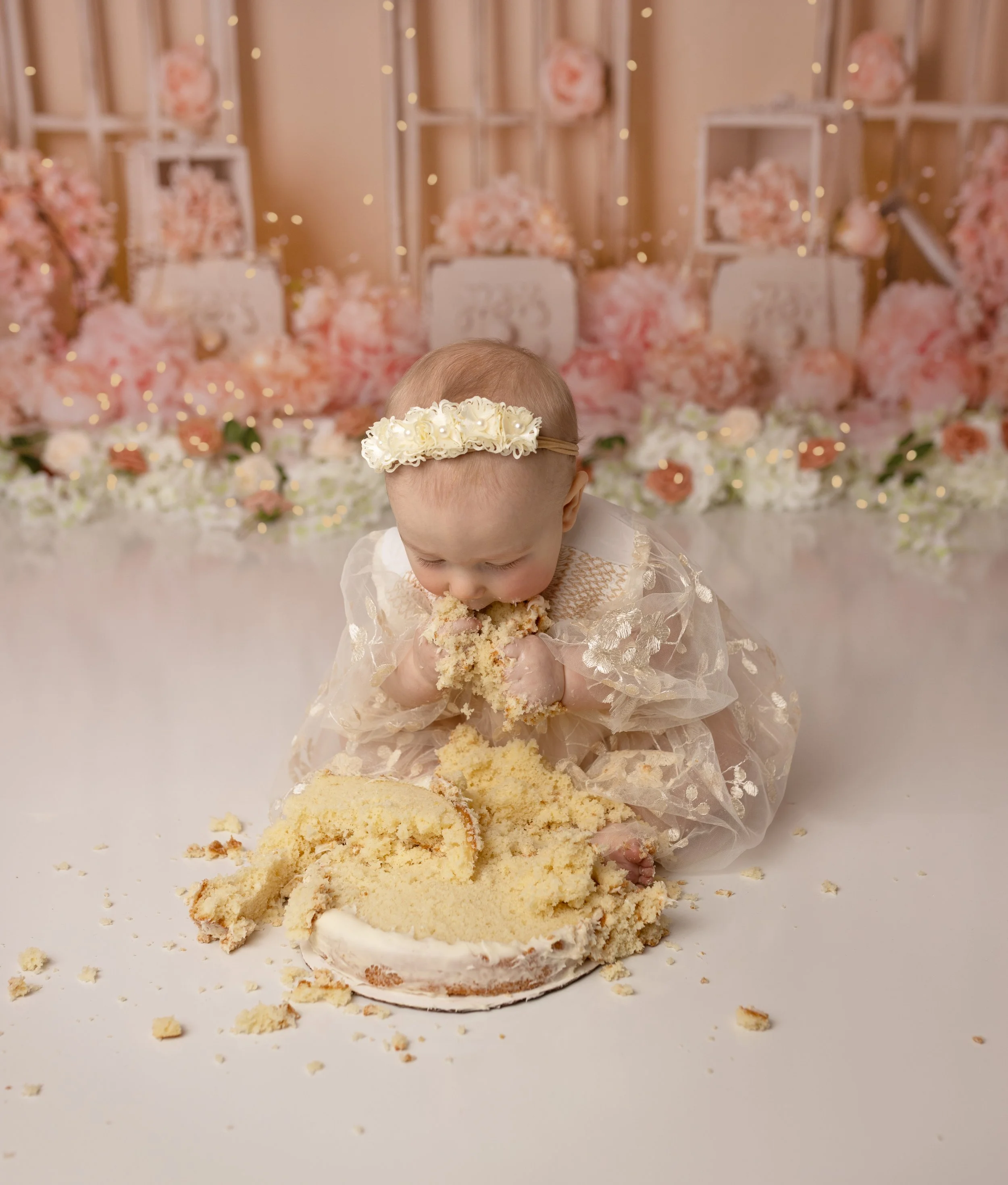 A baby girl wearing a cream dress and floral headband eating a cake during a celebration with a pink and white floral backdrop.
