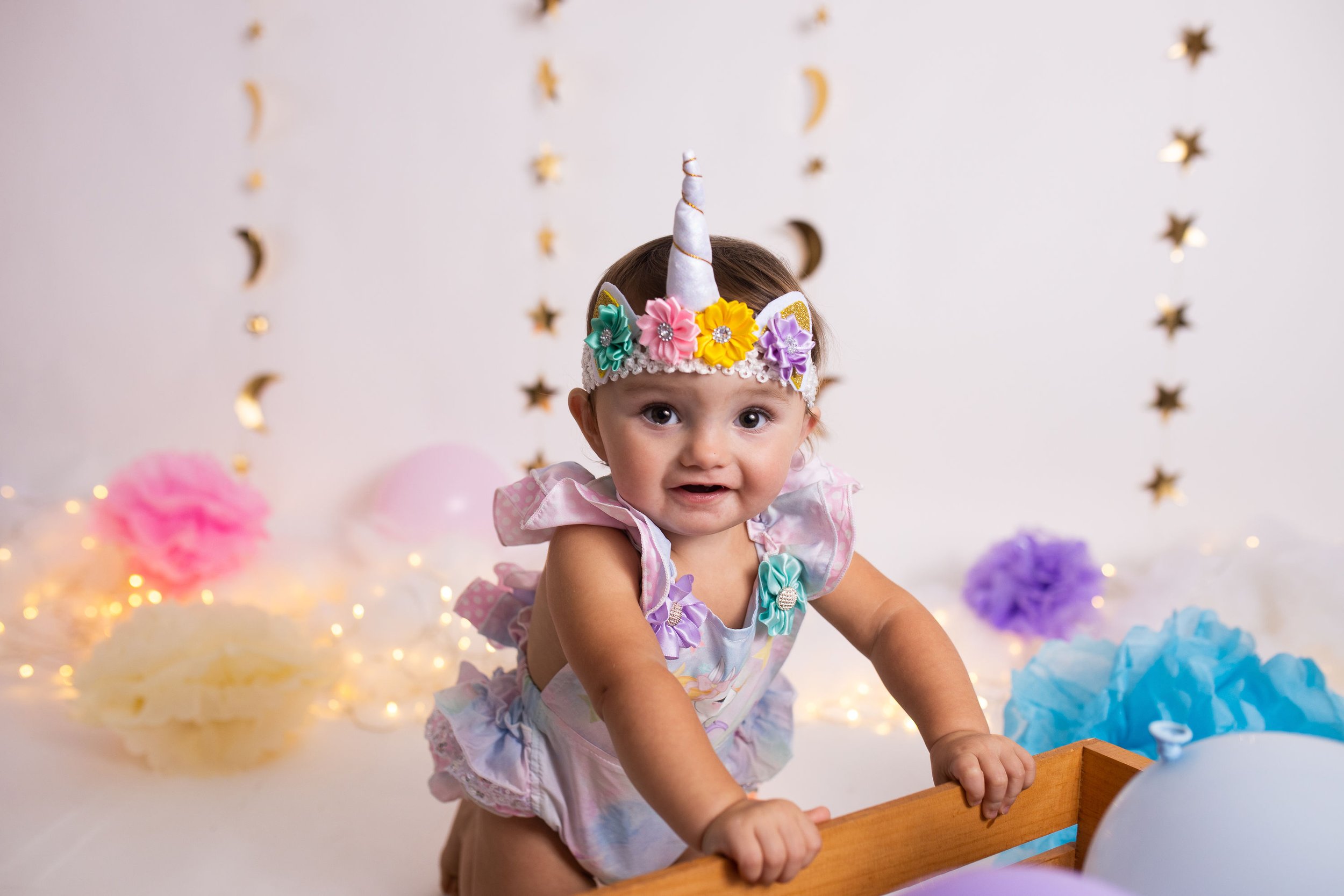 Baby wearing a unicorn headband and floral romper, surrounded by colorful decorations and lights.