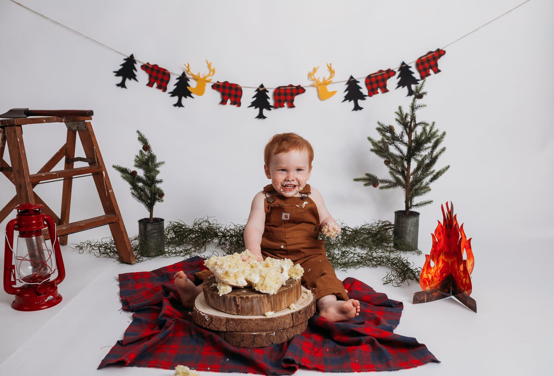 Smiling toddler in brown overalls seated on a red and black plaid blanket, surrounded by woodland-themed props such as small trees, a paper campfire, and a red lantern, with a garland of bears and trees in the background. The child is playfully cover
