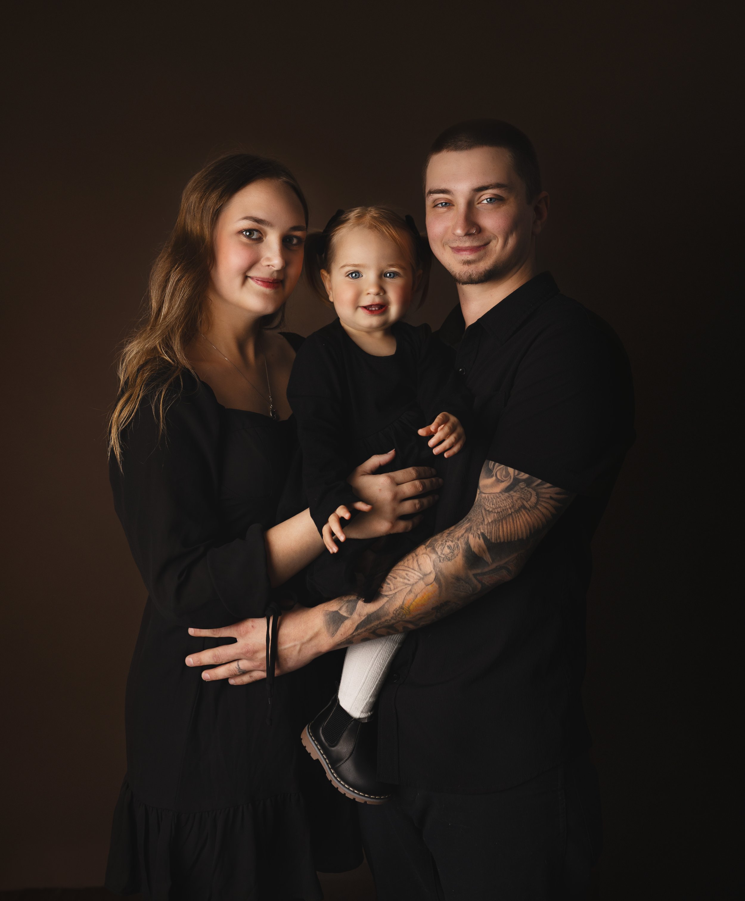 A family of three: mother, father, and young daughter, all dressed in black, standing against a dark background, smiling and posing for a portrait.
