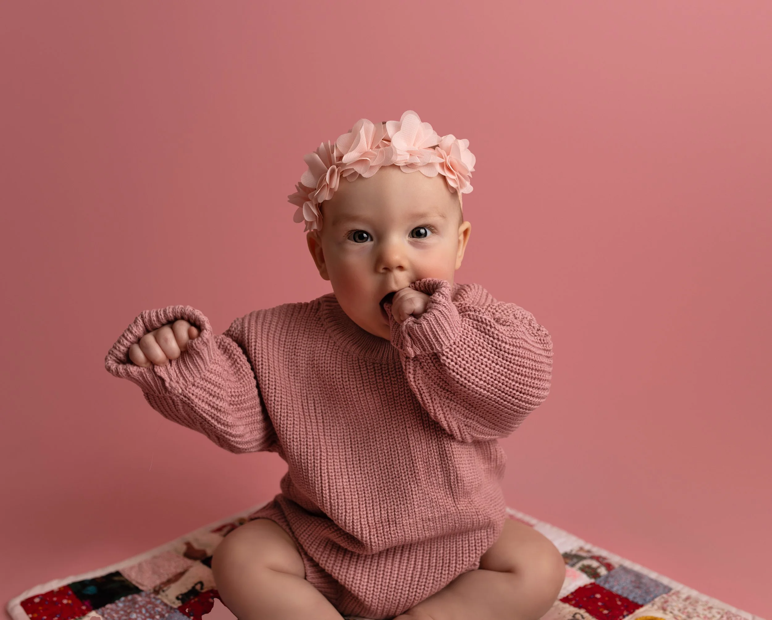 A baby girl with a pink flower headband, wearing a pink knit sweater, sitting on a patchwork quilt against a pink background, with one hand in her mouth and the other raised in a fist.