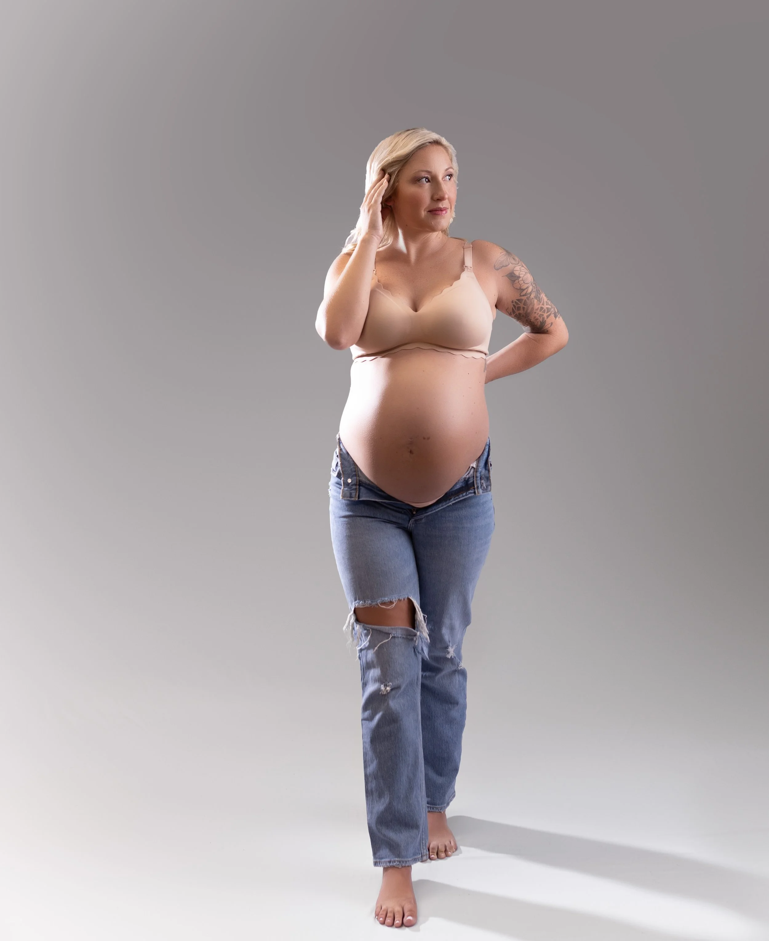 Pregnant woman with blonde hair, wearing a beige bra and ripped jeans, standing barefoot against a plain gray background, with one hand on her head and the other behind her backLouisville ky maternity photographer
